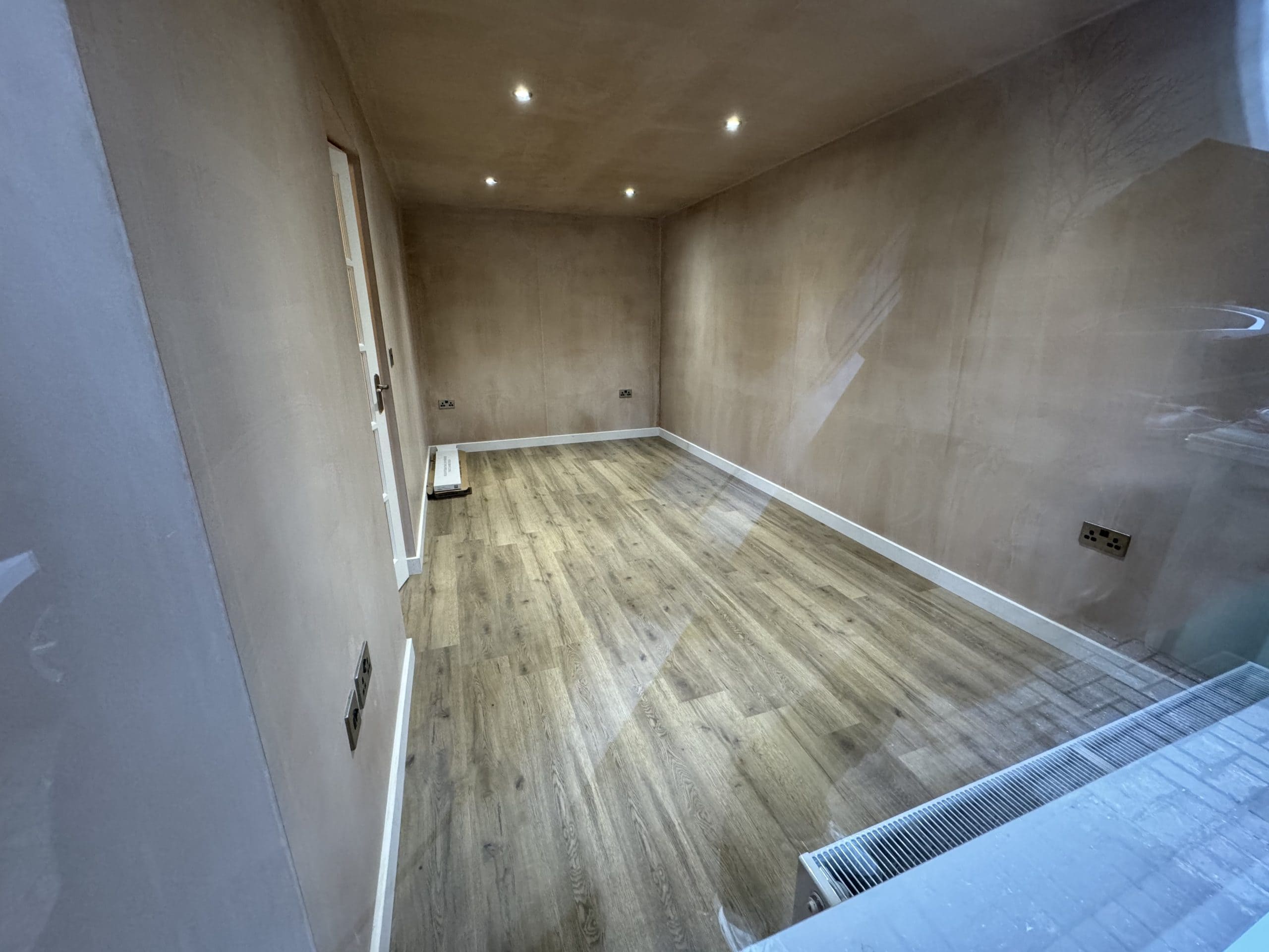 A small, empty room with newly plastered walls, wood-effect flooring, and recessed ceiling lights—perfect for a kitchen or bathroom showroom installation. Features include a radiator under the window, a step, and accessible electrical sockets.