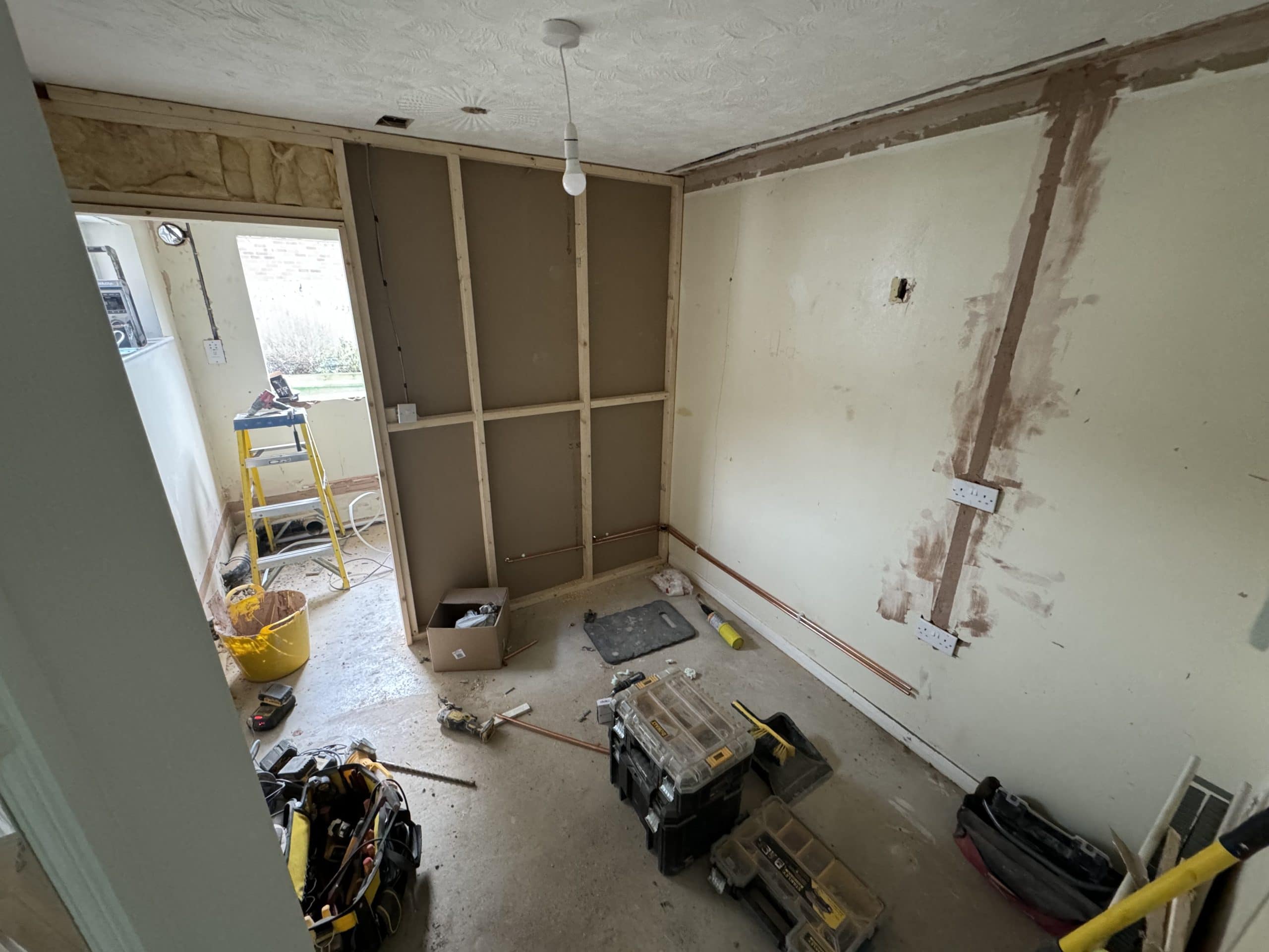 A partially renovated room with exposed wooden wall framing, construction tools, toolboxes, and building materials scattered on the unfinished floor. Perfect for planning a kitchen and bathroom showroom or installation project; patchwork is visible on the walls.
