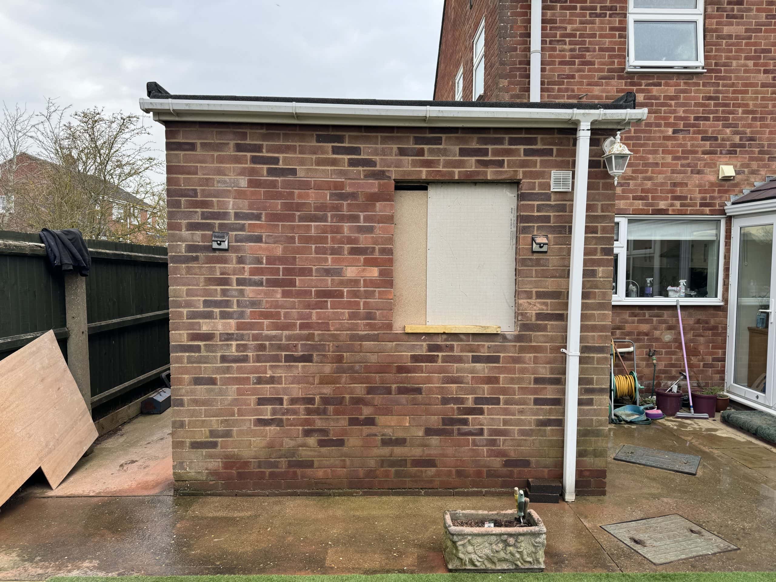 A small brick outbuilding with a boarded-up window stands next to a house, hinting at potential kitchen and bathroom showroom installation possibilities. A black gutter, drainpipe, stone planter, and paved ground complete the design beside a wooden fence.