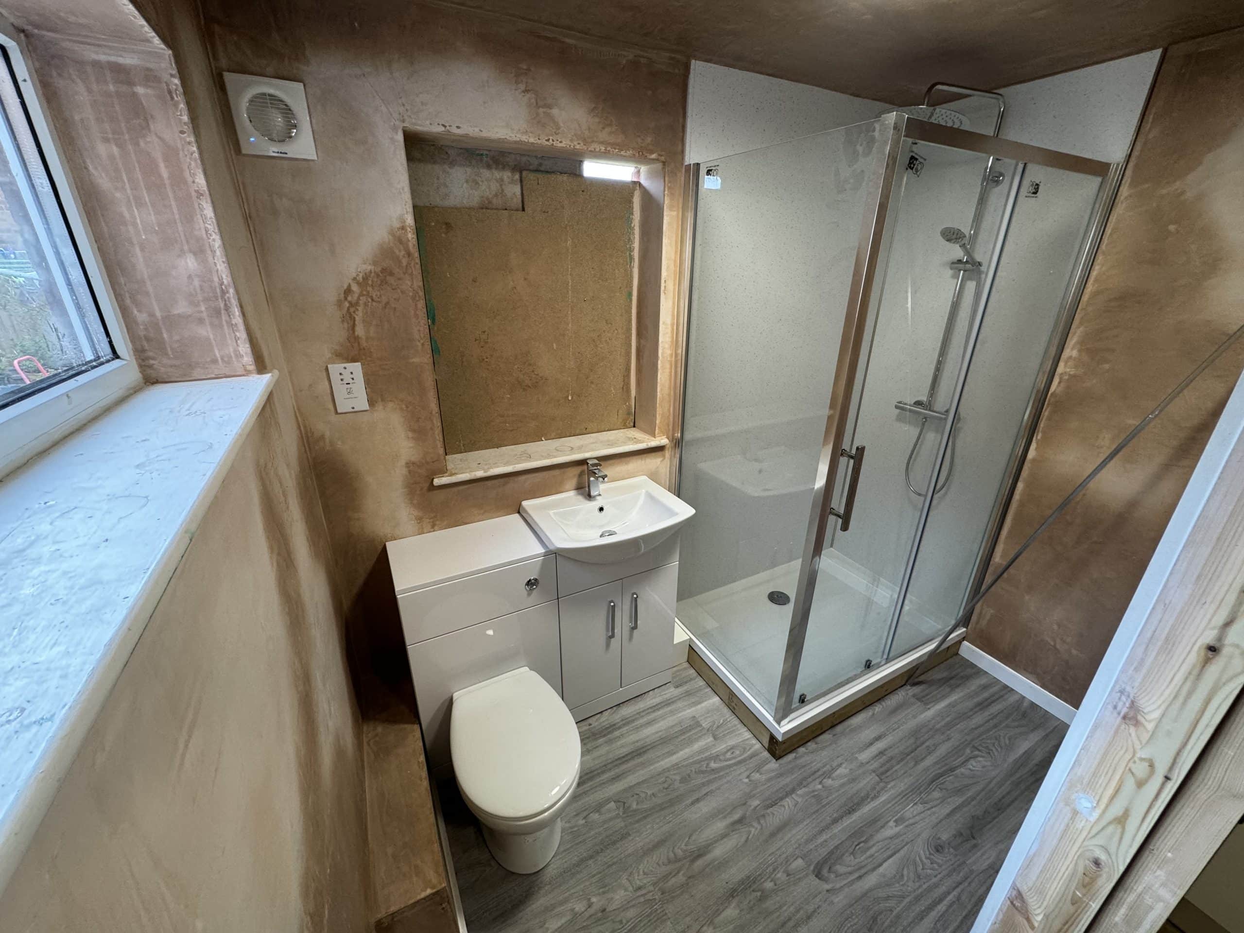 Newly renovated bathroom with unfinished plaster walls, featuring a toilet, a sink with cupboard, and a glass shower enclosure—perfect inspiration for your next kitchen and bathroom showroom design or installation project. Wood-effect vinyl flooring included.