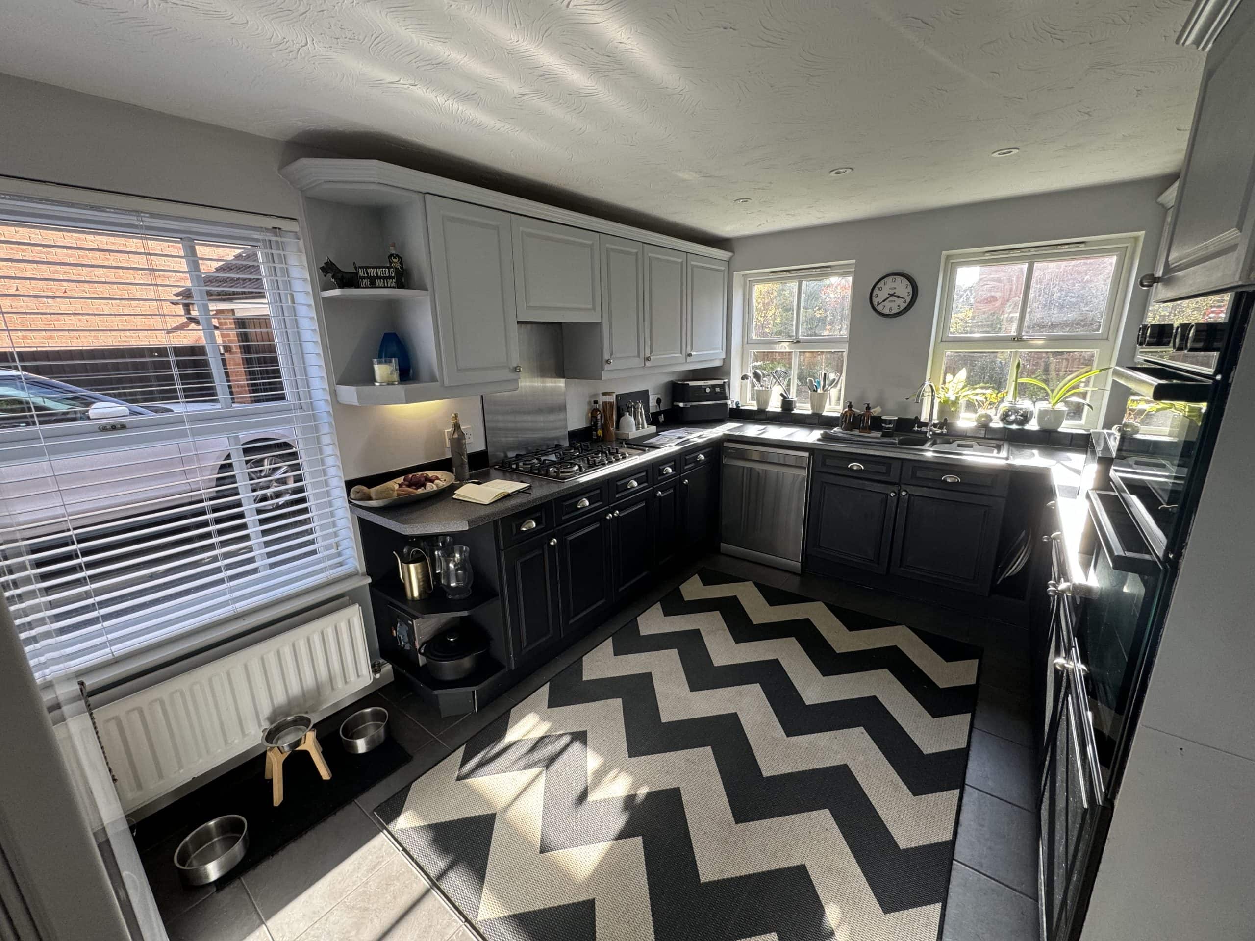 A modern kitchen and bathroom showroom-inspired design with grey cupboards, black lower units, and a black and white zigzag rug. Sunlight streams through two large windows above the sink, while dog bowls sit near a window with closed blinds.