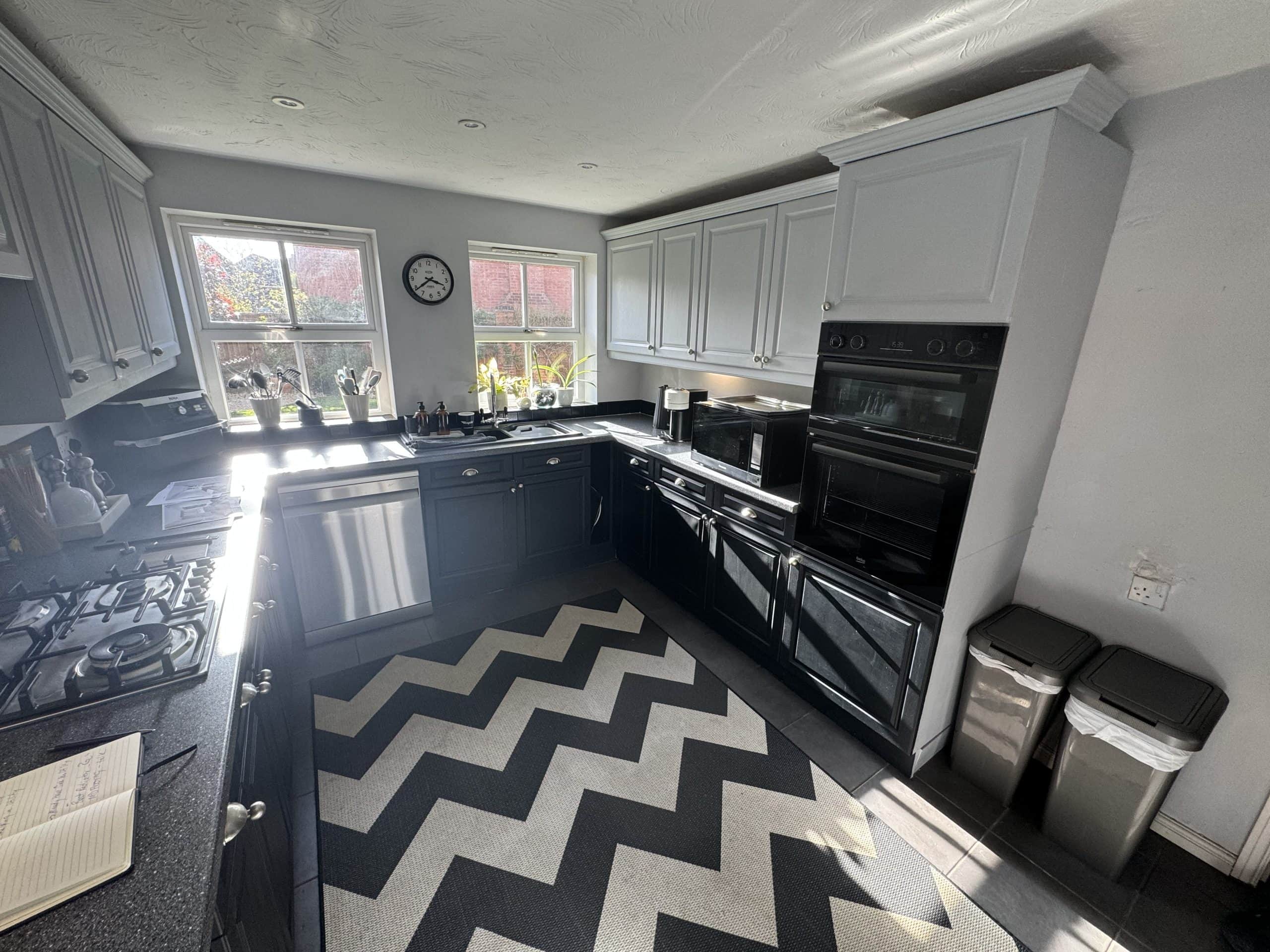 A modern kitchen design with dark cupboards, stainless steel appliances, two windows letting in sunlight, a zigzag-patterned rug, and two bins near the cooker. Potted plants line the windowsill, showcasing stylish installation supply inspiration.