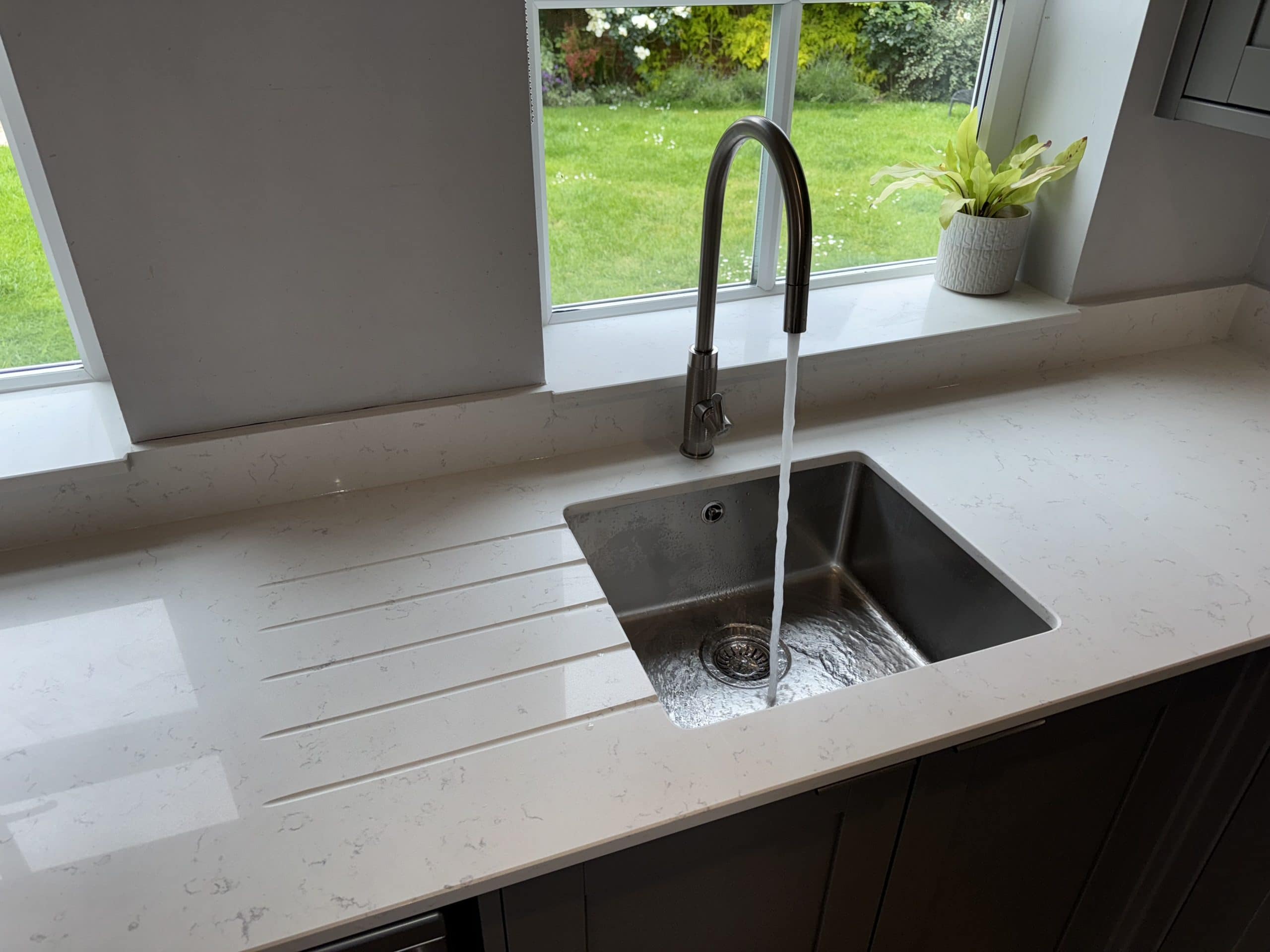 A stainless steel kitchen sink with running water is set in a white worktop beneath a window—a perfect example of showroom-worthy installation and design. A potted plant sits on the windowsill, overlooking the green lawn outside.