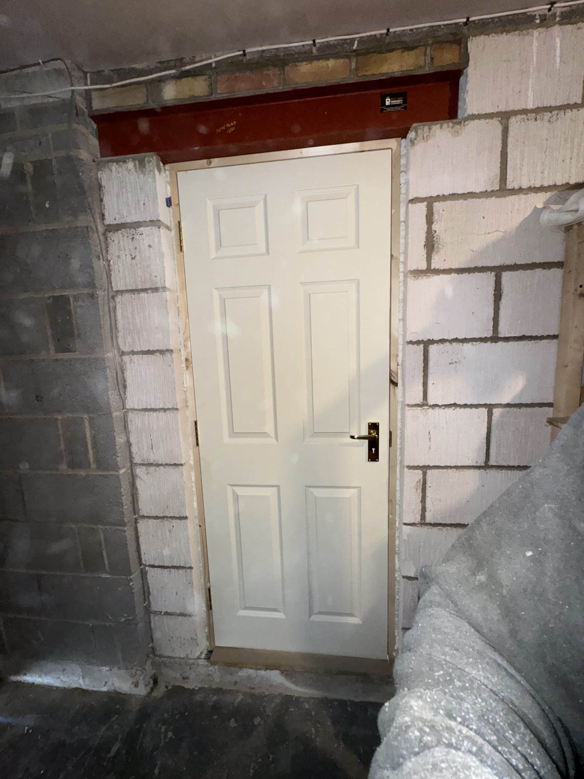 A white panelled door with a gold handle is set in a partially finished brick and concrete block wall, suggesting an area prepared for kitchen and bathroom showroom installation or design supply, with exposed bricks and mortar framing the entrance.