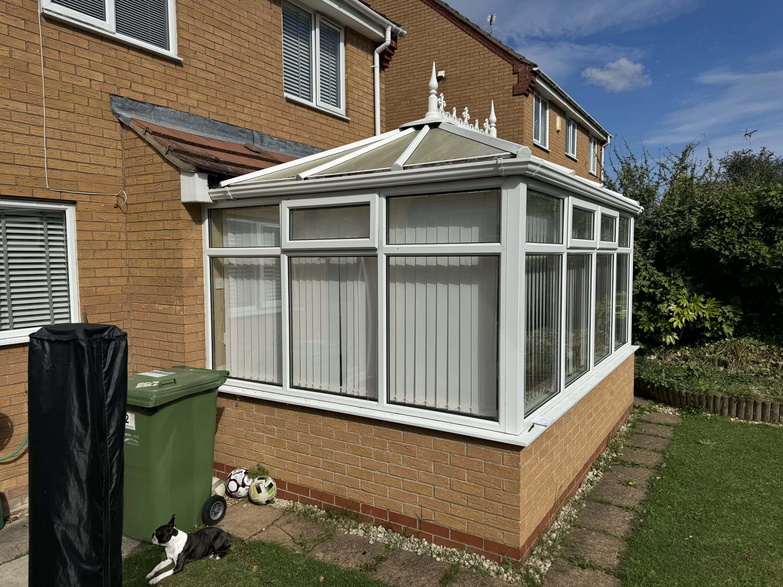 A white conservatory with vertical blinds attached to a brick house, next to a green bin and a black-and-white cat lying on the grass. Nearby are garden toys and a covered barbecue—perfect inspiration for any kitchen and bathroom showroom design. The sky is partly cloudy.