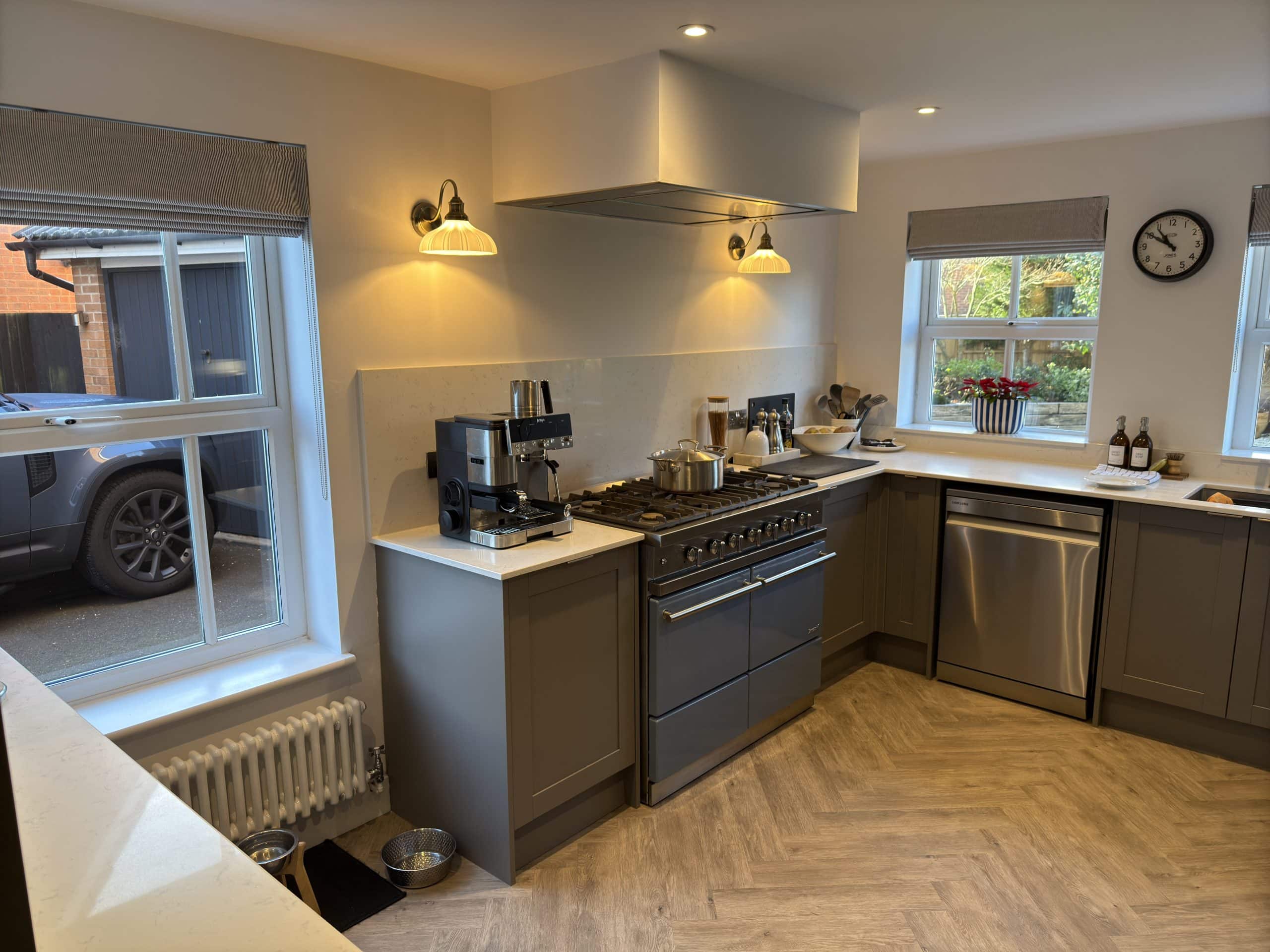 Modern kitchen and bathroom showroom-inspired design with grey cupboards, stainless steel cooker, dishwasher, and coffee machine. Large windows let in natural light. Clock on the wall, potted plant by the window, and pet bowls on the floor.