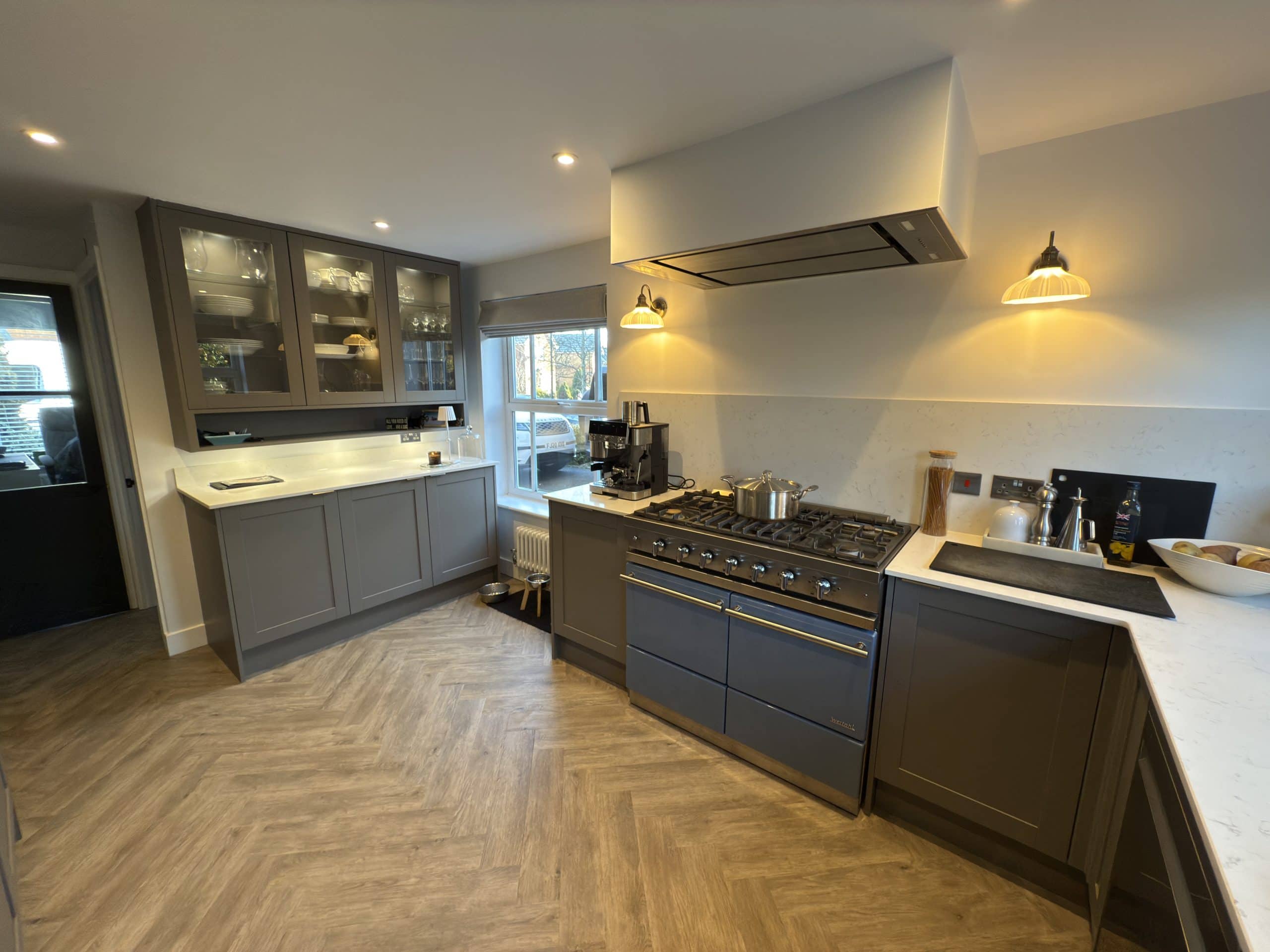 Modern kitchen and bathroom showroom design featuring grey cupboards, a large cooker, white worktops, glass-fronted cabinets, wooden herringbone flooring, and under-cupboard lighting; window and door let in natural light.