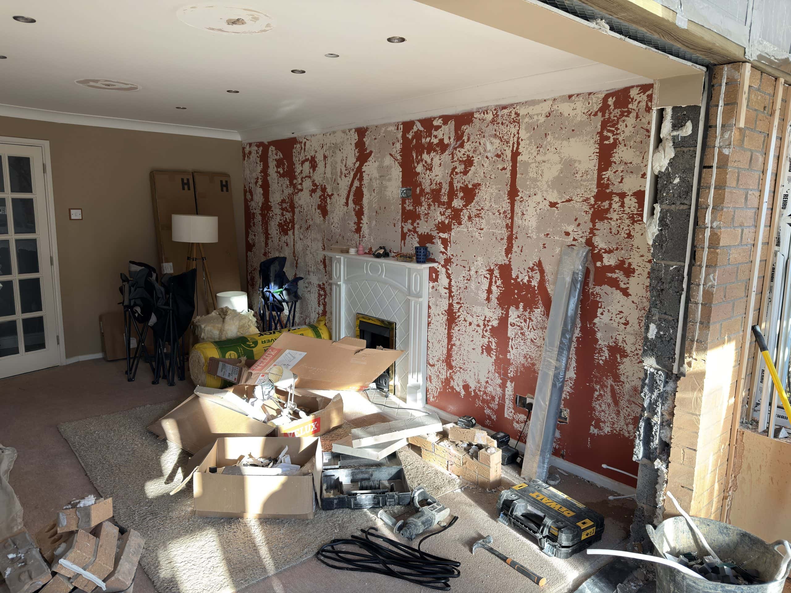 A living room undergoing renovation with exposed plaster on a red wall, tools and boxes from a kitchen and bathroom showroom scattered on the floor, a fireplace, and an unfinished opening in the brick wall on the right.