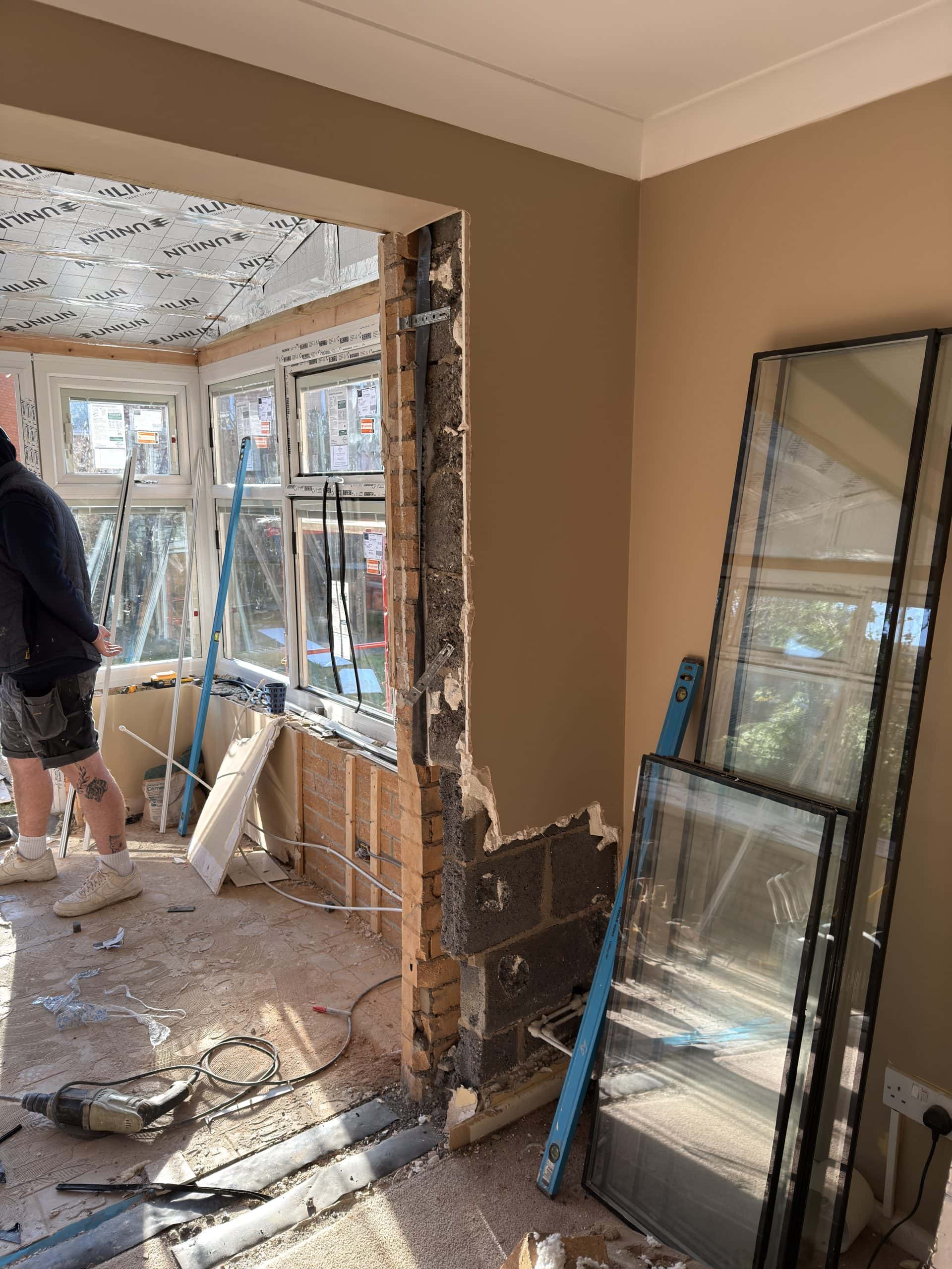 A partially demolished interior wall in a house undergoing renovation, with exposed bricks and insulation visible. Kitchen and bathroom showroom design elements and installation tools are scattered about. A person stands near a large window area.