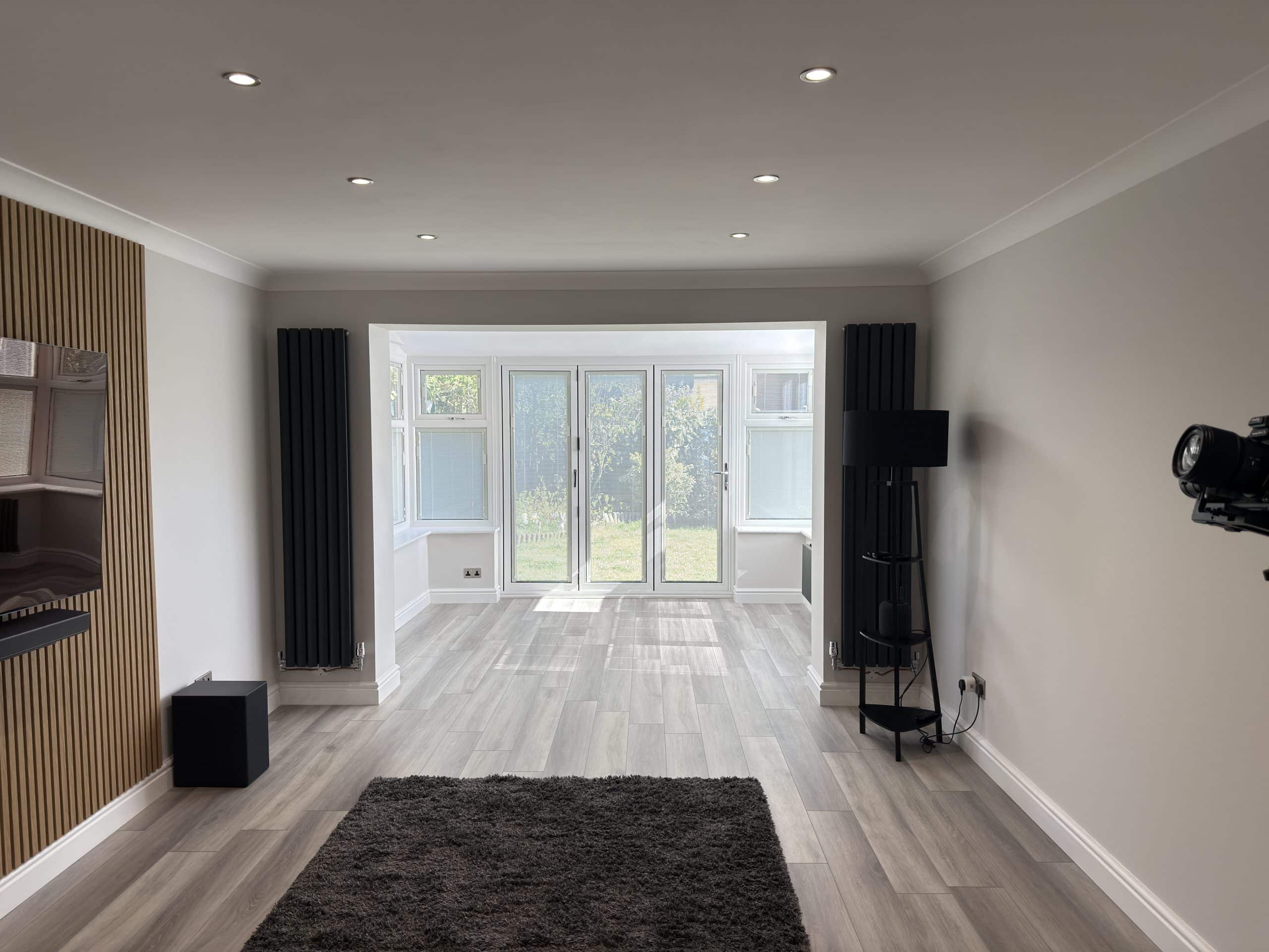 A modern, unfurnished lounge with light grey walls, wooden flooring, and recessed ceiling lights. Perfect for showcasing kitchen and bathroom showroom installation supply design ideas, it features large windows and glass doors opening to a green garden view.