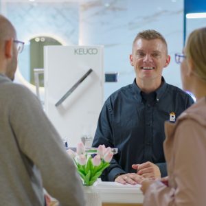 A smiling sales assistant stands behind a counter, engaging with two customers. Pink tulips in a vase and a KEUCO display enhance the bright, modern kitchen and bathroom showroom design.