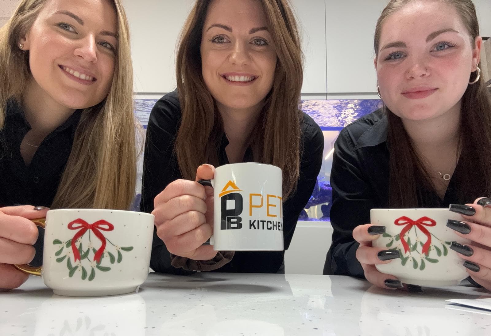 Three women smiling and holding mugs while sitting at a white worktop. Two mugs have a festive design with a red bow and green leaves, and the centre mug has the logo DB PET KITCHEN.