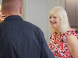 A smiling older woman in a pink floral dress talks with a man in a dark shirt indoors. Another person is partially visible on the left side of the image.