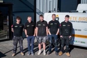 Five men wearing black company shirts stand side by side, smiling, in front of a white van with business signage on a sunny day outside a modern industrial building.