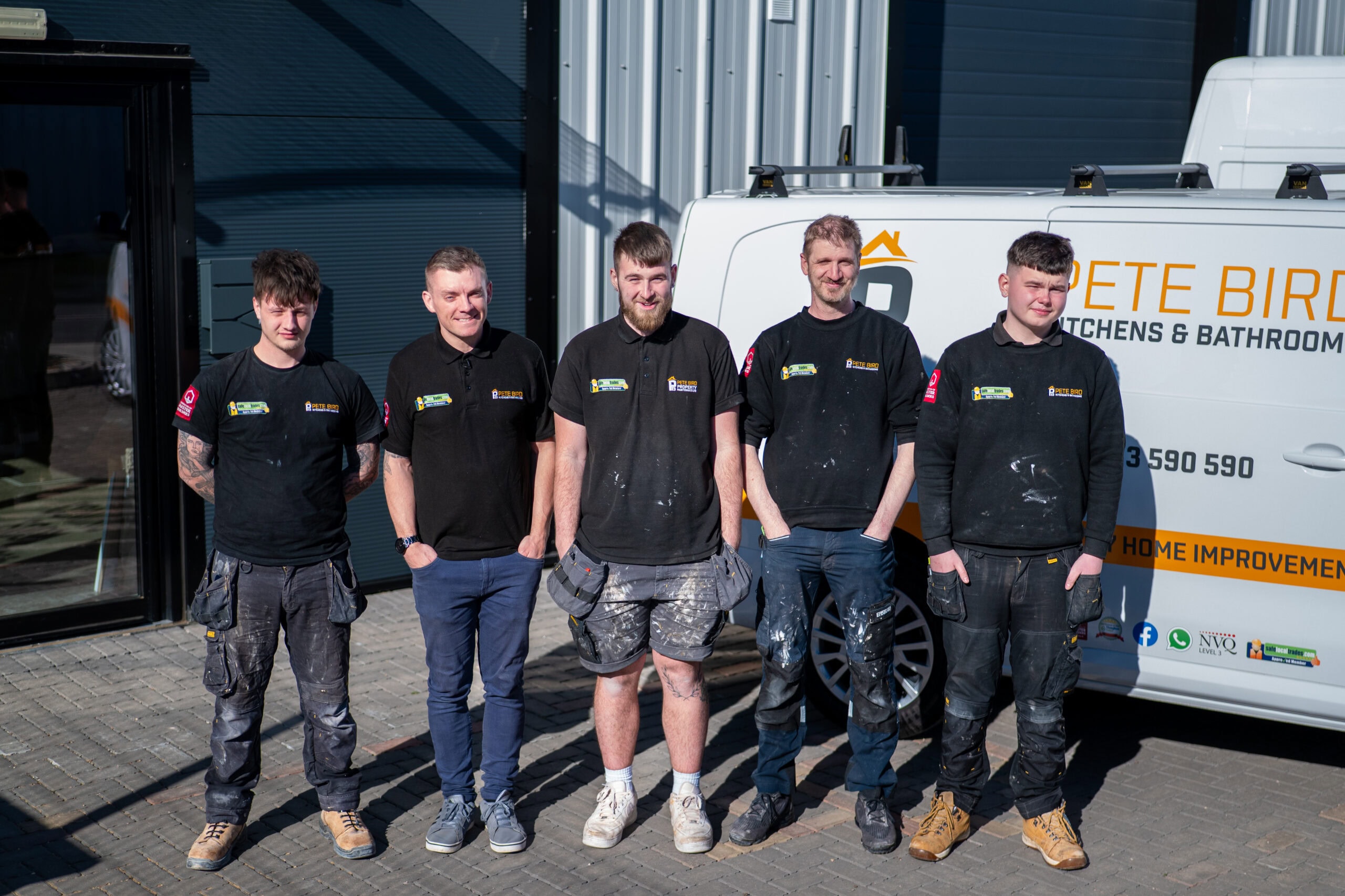 Five men wearing black company shirts stand side by side, smiling, in front of a white van with business signage on a sunny day outside a modern industrial building.
