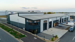 Aerial view of a modern industrial building with multiple units, including Pete Bird Construction, wide entrances, a parking area with vehicles, and surrounding roads during daytime.