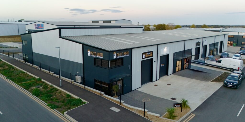 A modern industrial building with grey and white cladding, large windows, and Pete Bird Construction signs. Several parked vehicles and a paved pavement surround the structure. The sky is partly cloudy.