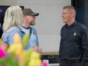 Three adults are talking indoors. A man in a black shirt speaks to a man wearing a cap and glasses, who stands beside a woman with blonde hair. Colourful blurred flowers are in the foreground.