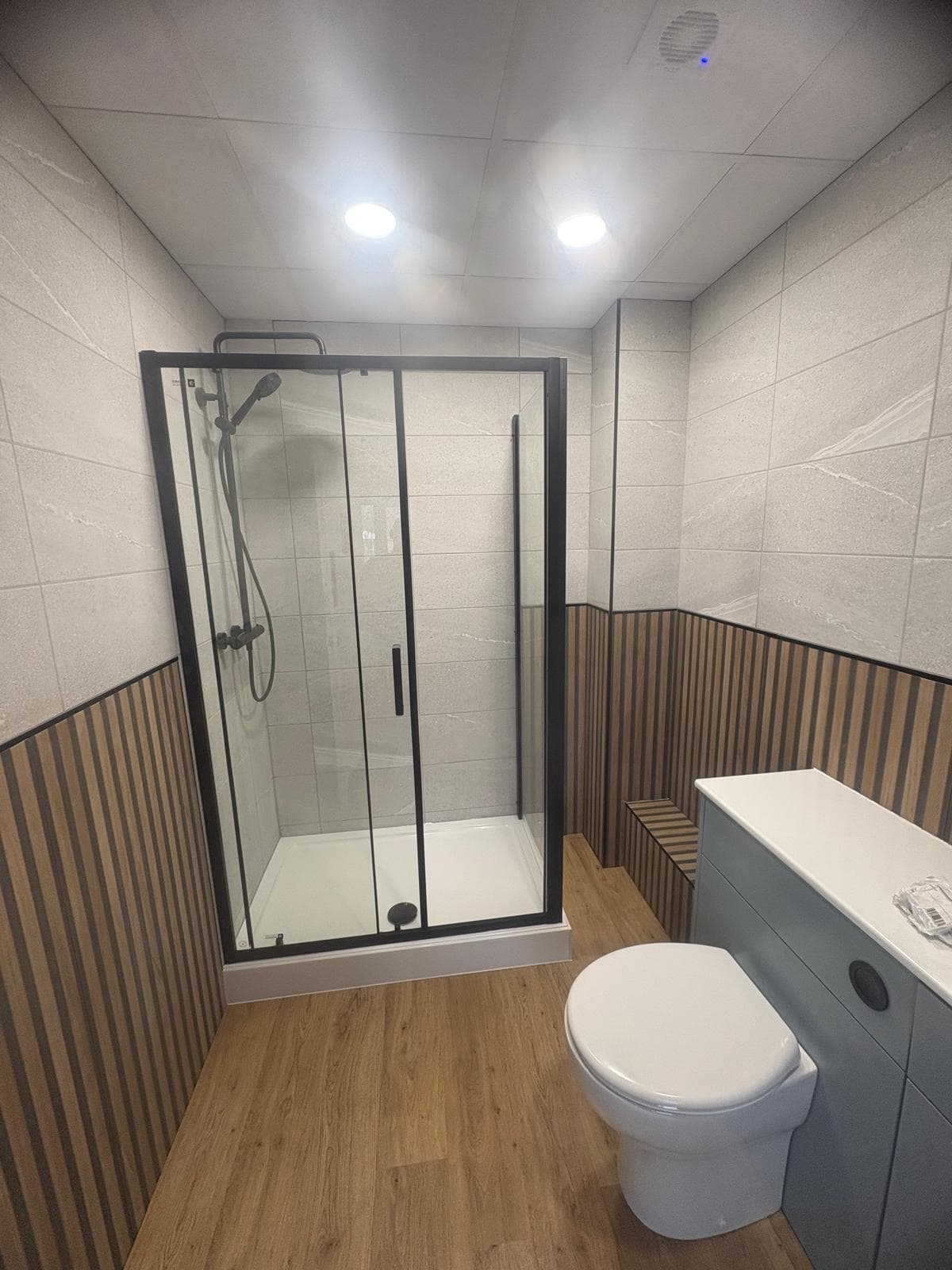 Modern bathroom with a glass shower enclosure, rainfall showerhead, white toilet, and light blue vanity unit. Walls have light tiles above wooden slats and the floor is wood-effect. Ceiling lights illuminate the space.