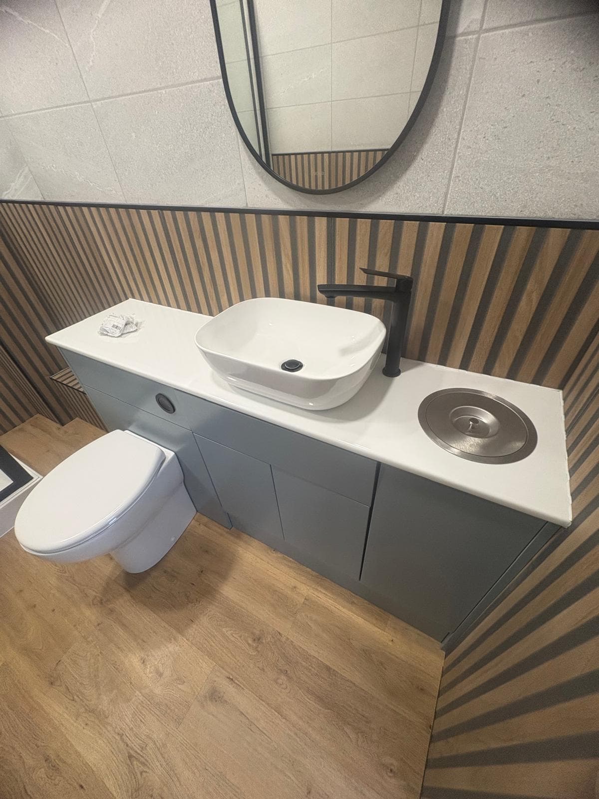 A modern bathroom with a white countertop, vessel basin, black tap, and circular waste bin. The vanity has blue cupboards, a wall-mounted toilet, and wood-patterned and tiled wall panels. A round mirror is above the basin.
