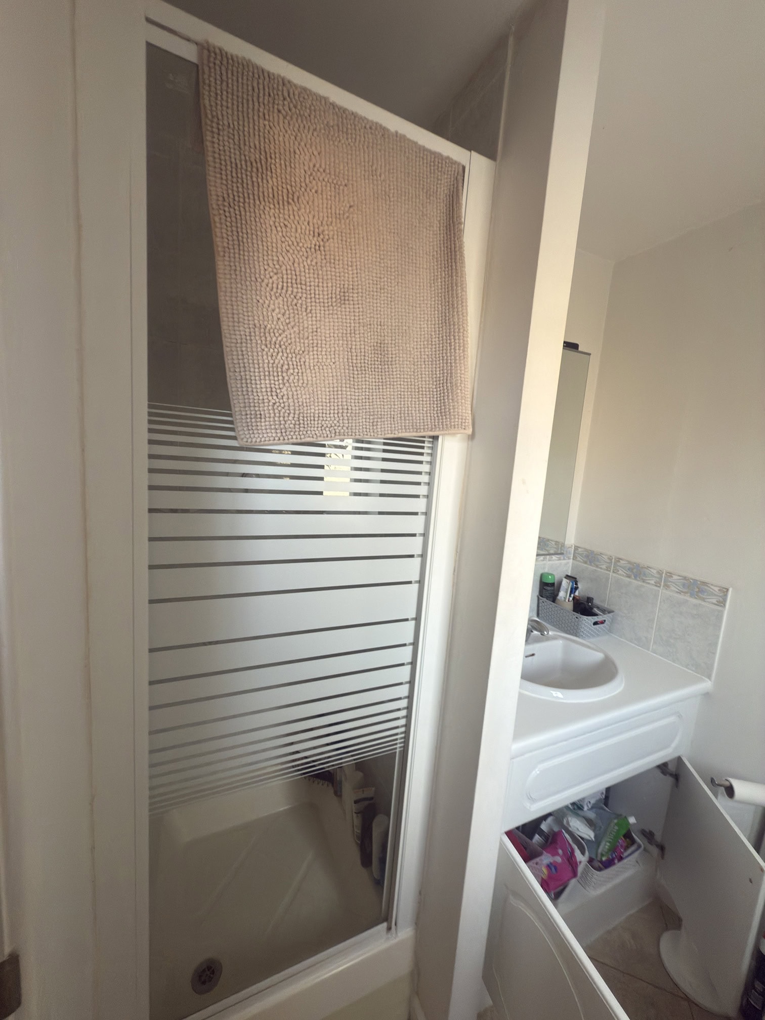 A small bathroom with a glass shower door featuring horizontal frosted stripes, a beige towel hanging over the top, and a sink with an open cupboard door below it. Toiletries are visible on the counter.