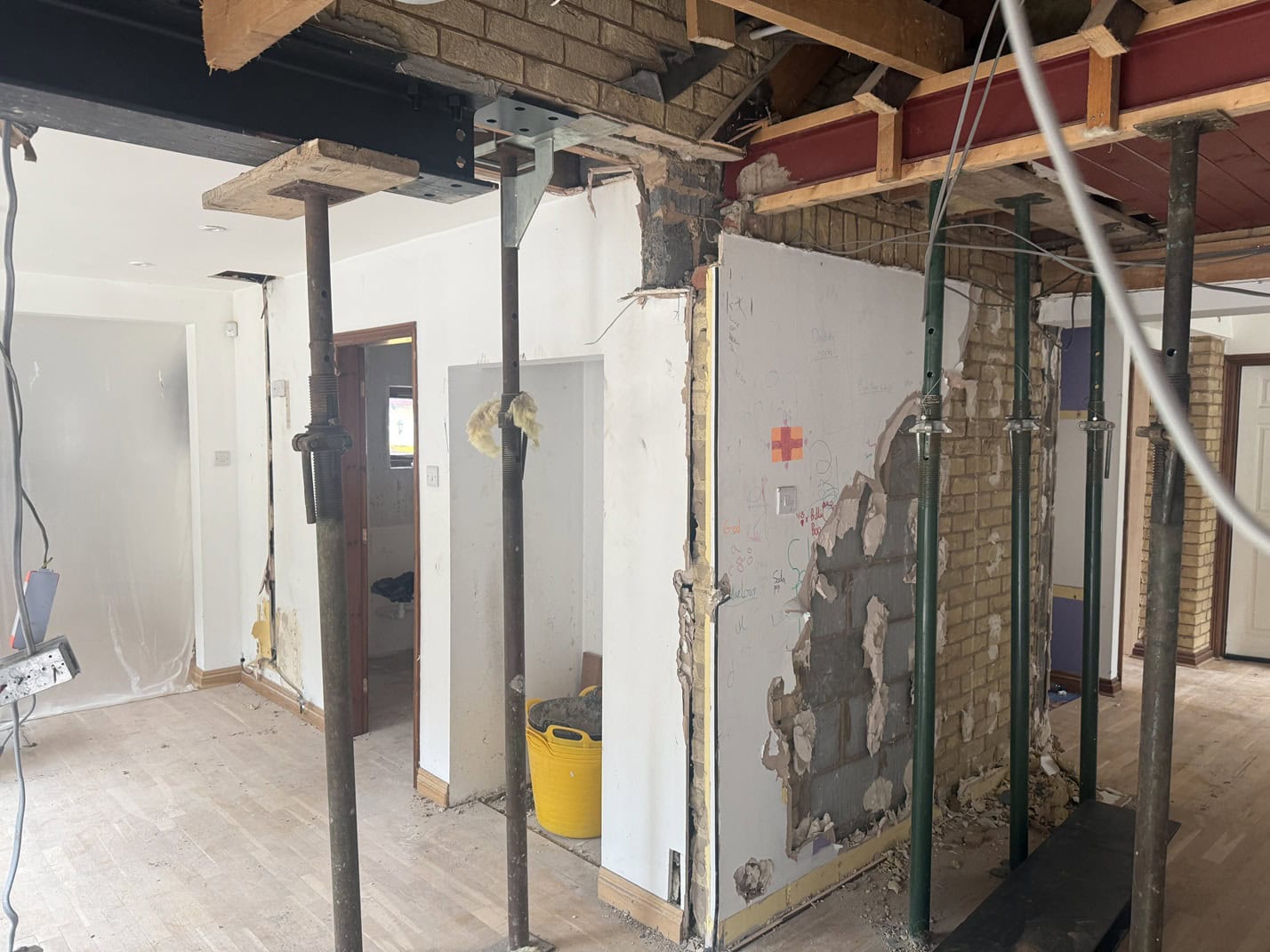 A partially demolished interior wall supported by metal beams and poles, with exposed brickwork and insulation. Construction materials and debris are scattered on the floor of the refurbishment site.