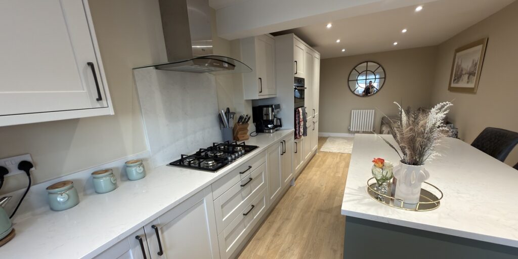 Modern kitchen with white cupboards, gas cooker, and countertop appliances. Light wood floor, neutral walls, decorative vases, and a tray with dried flowers on the island. Round mirror and framed picture in the dining area.