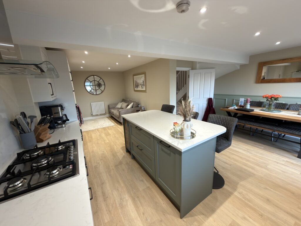 Open-plan kitchen and living area with a central island, gas hob, grey cupboards, wooden floors, dining table with bench seating, a sofa, large clock on the wall, and neutral décor.