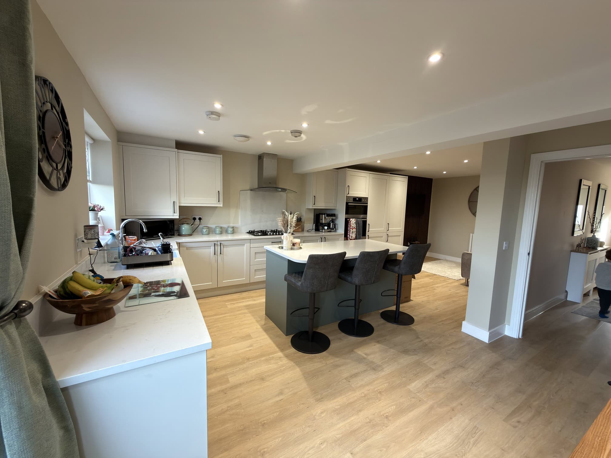 Modern kitchen with white cupboards, an island with three grey barstools, light wood flooring, and built-in appliances. A bowl of fruit and a book are on the worktop, and a person is partially visible on the right.