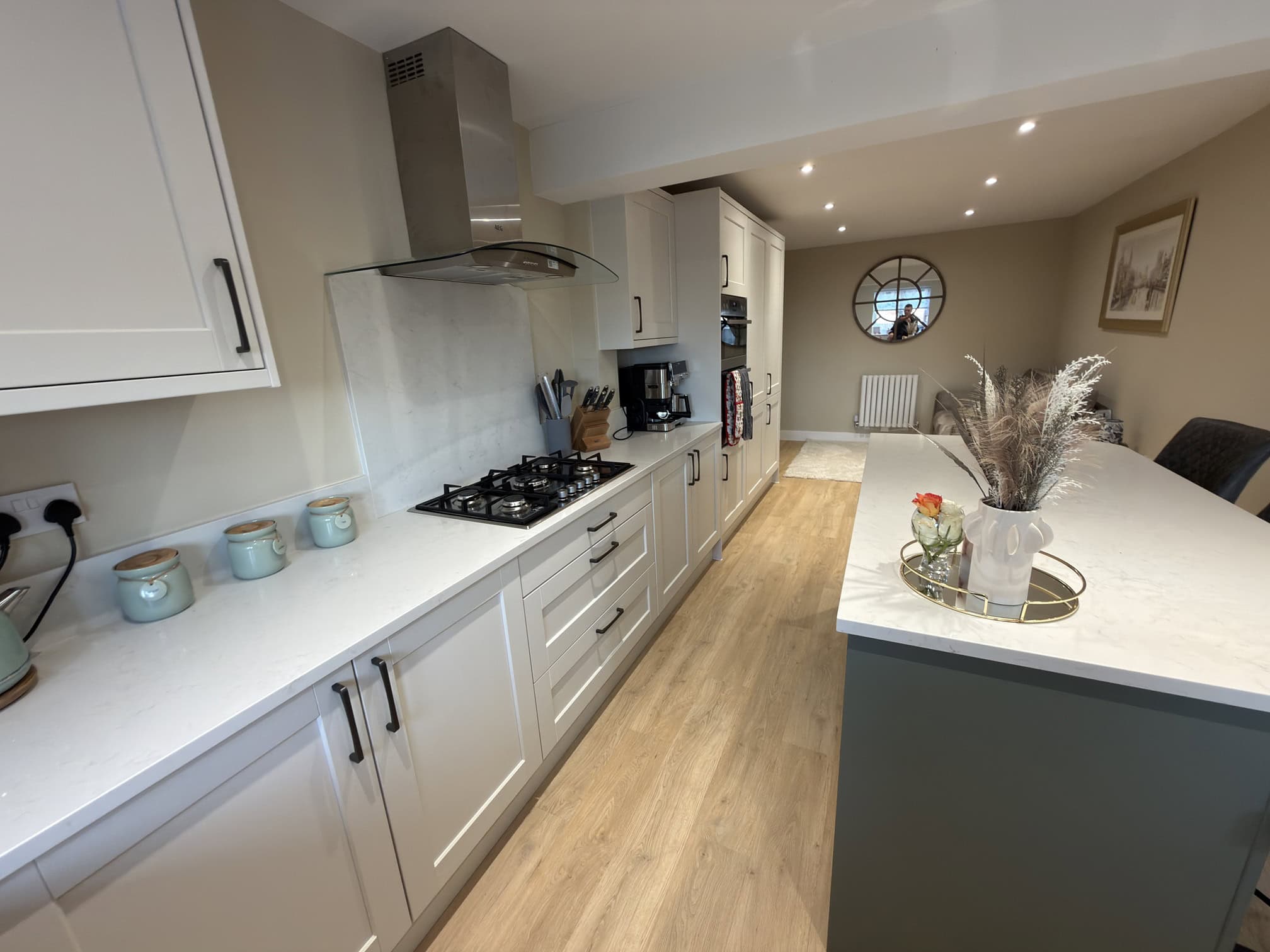 Modern kitchen with white cupboards, gas cooker, wooden flooring, and a large island with a decorative tray holding a vase of dried flowers. Neutral walls, recessed lighting, and a circular wall mirror create a bright, cosy atmosphere.