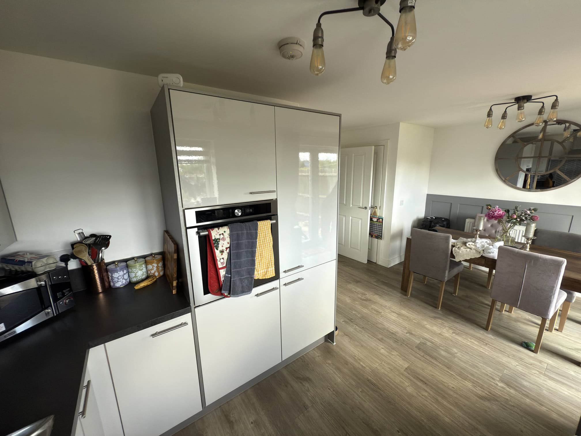 Modern kitchen and dining area with light wood flooring, white cupboards, built-in oven, and colourful towels hanging. Dining table set for four with flowers, mirror, and light fittings overhead.