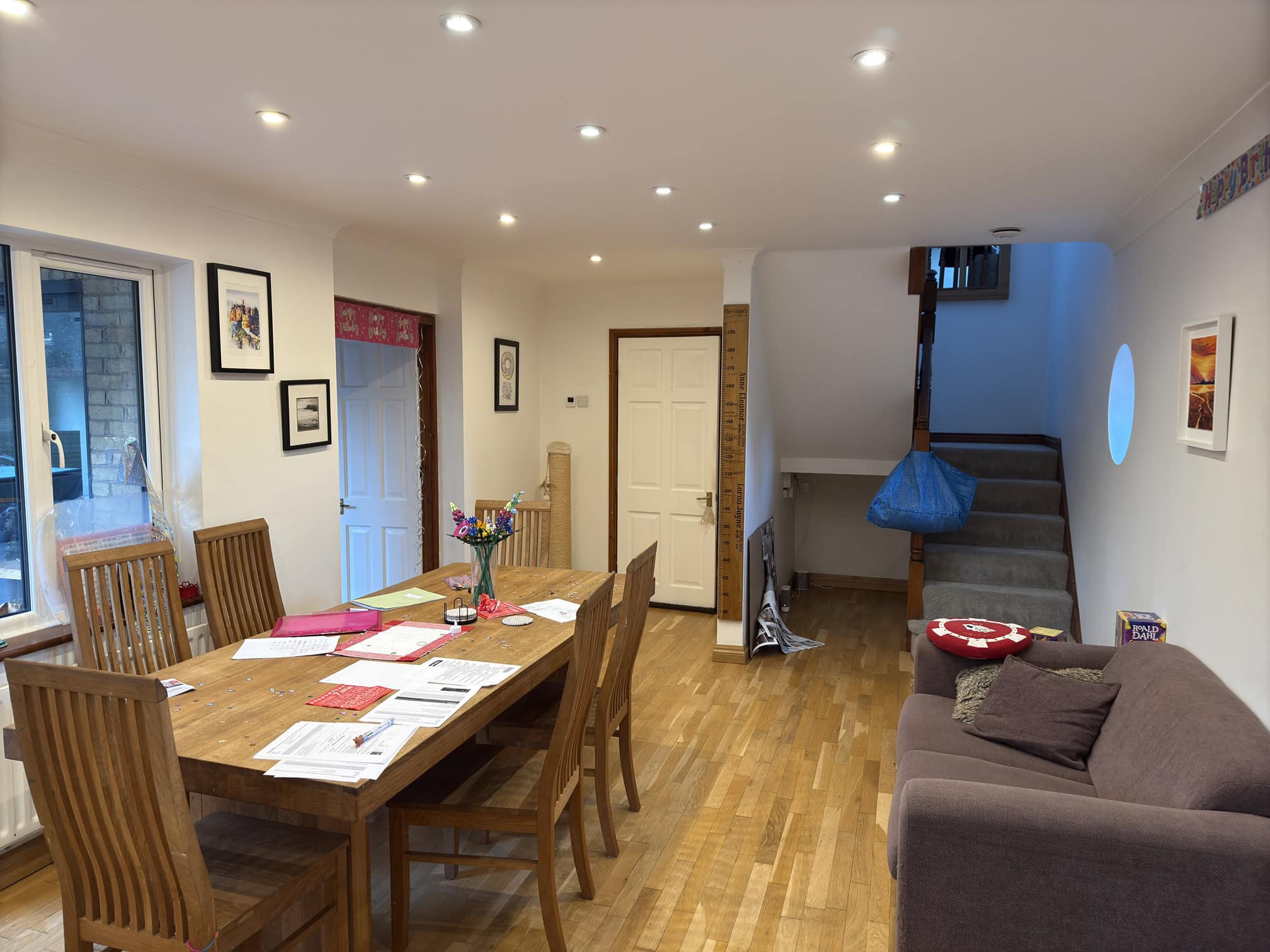 A cosy dining and living room with wooden floors, a table and chairs, papers scattered on the table, a brown sofa, stairway, artwork on the walls, and bright ceiling lights.