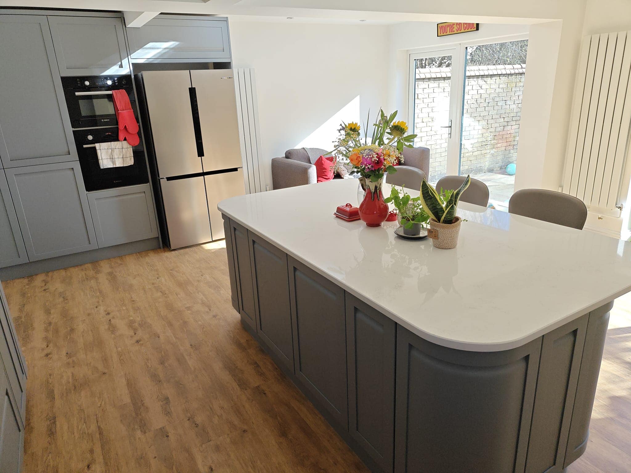 A modern kitchen with grey cupboards, wooden flooring, a large white island with flowers and plants on top, a stainless steel fridge, built-in oven, and a sitting area with chairs and a sofa by glass doors leading outside.