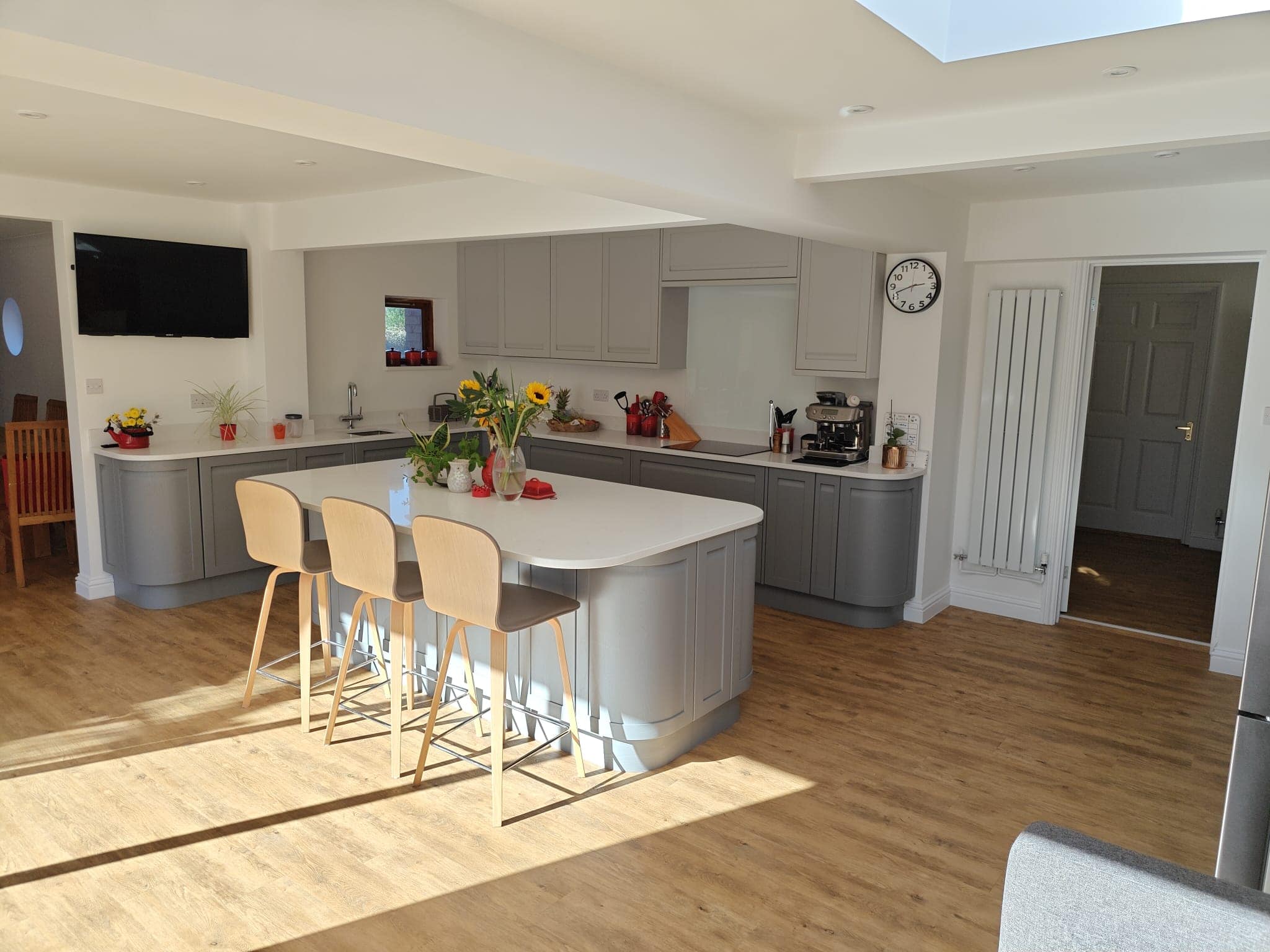 Modern kitchen with light grey cabinets, a large island with three beige bar stools, wooden floor, sunflowers on the worktop, wall clock, coffee machine, and sunlight streaming in through a skylight.