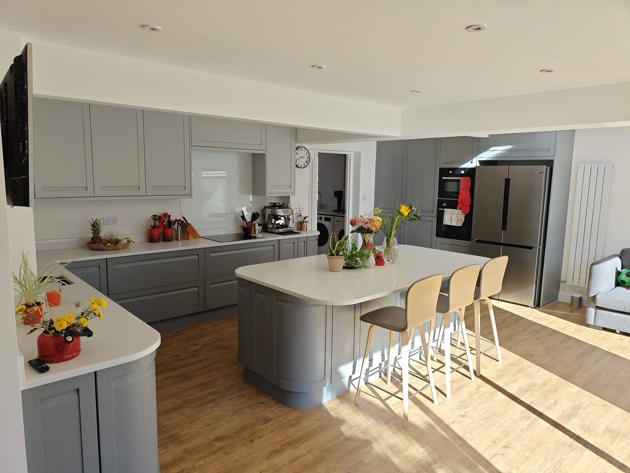Modern kitchen with grey cupboards, white worktops, and wooden flooring. There are three beige bar stools at an island, fresh flowers, fruit, and kitchen appliances visible, with sunlight streaming in through large windows.