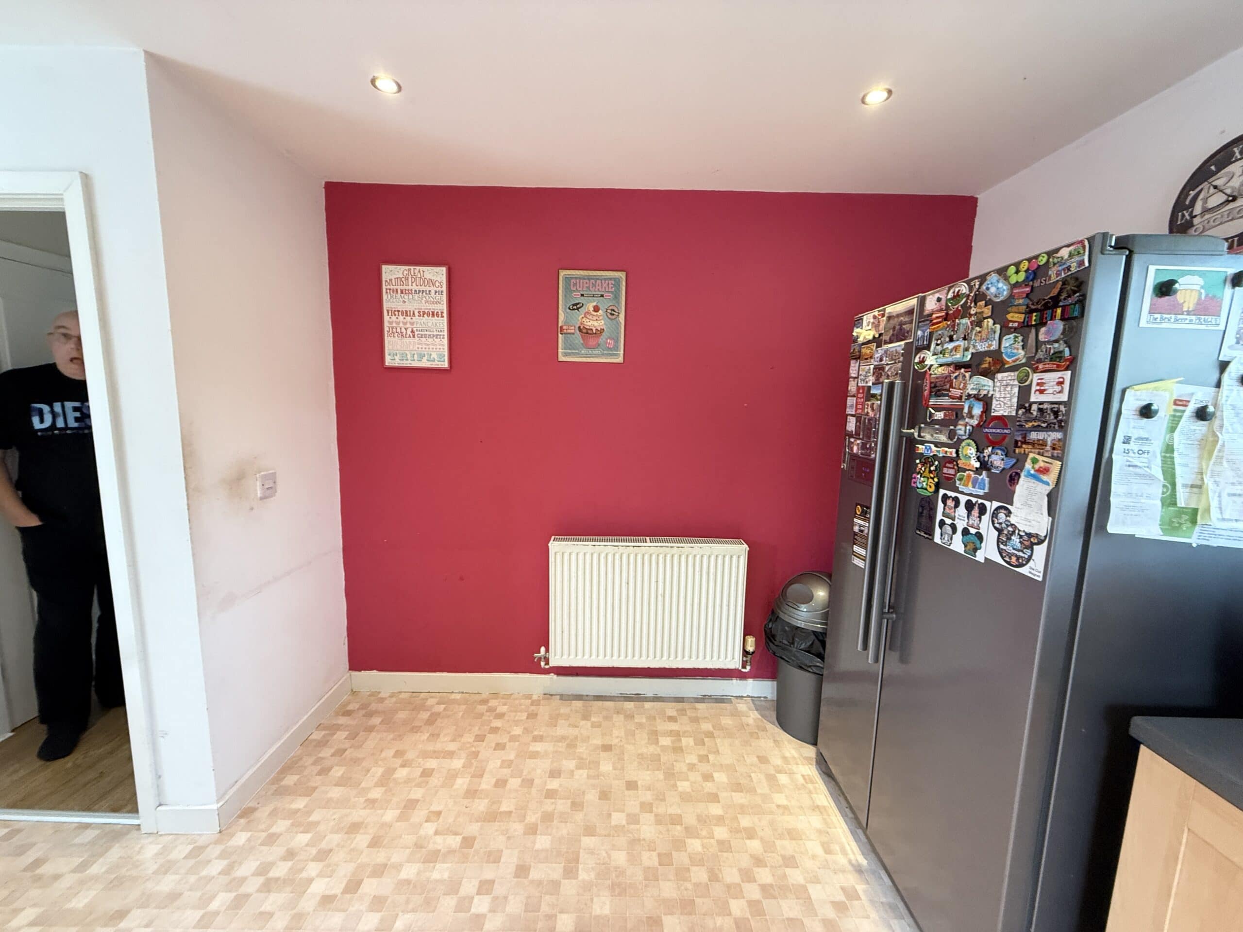 A kitchen with a pink feature wall, radiator, and two small posters. A grey fridge covered in magnets stands on the right, with a rubbish bin beside it. A man stands partially visible in a doorway on the left.