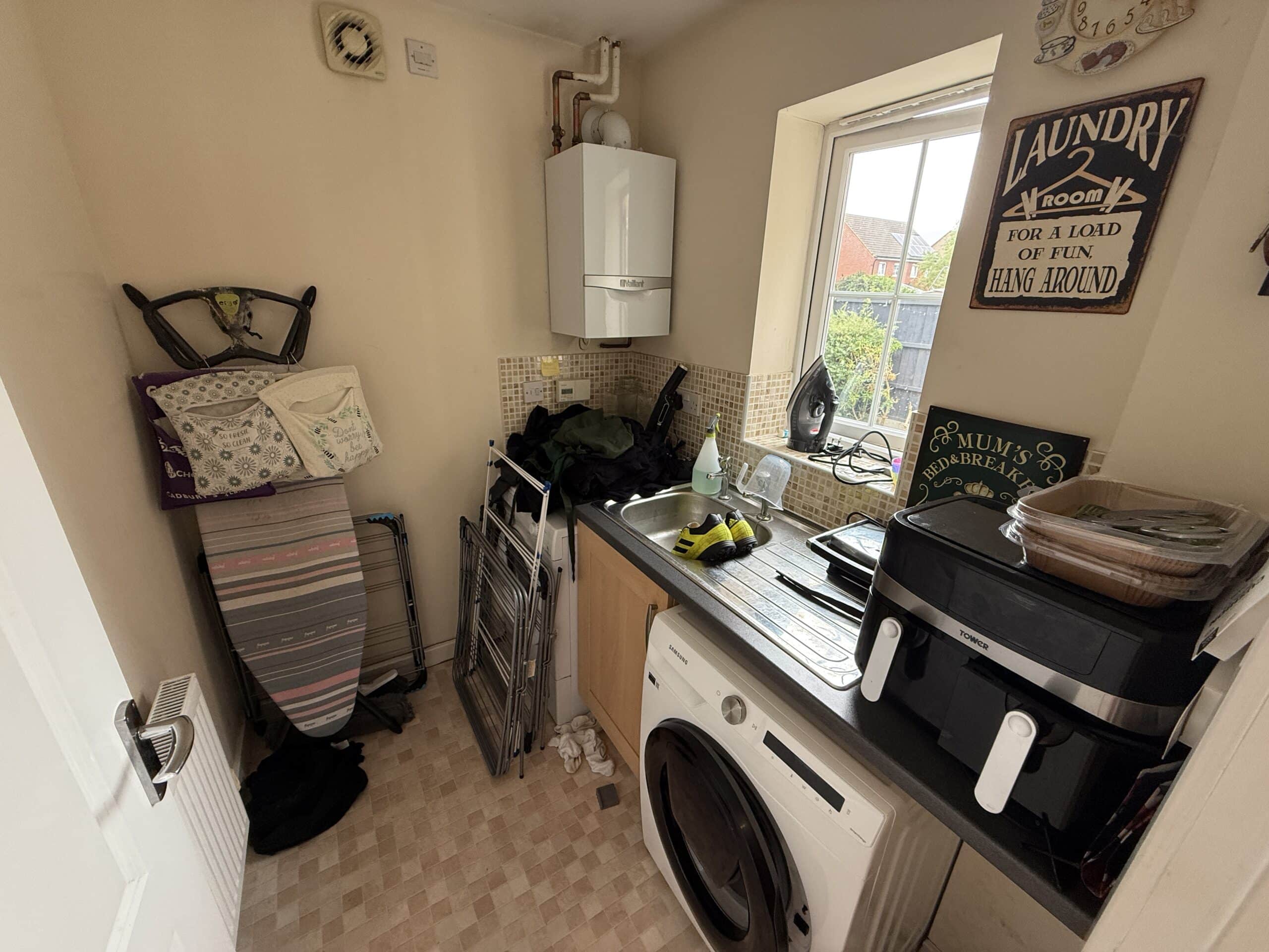 A small laundry room with a washing machine, ironing board, folded laundry, various household items on the worktop, and a window letting in natural light. A “Laundry Room” sign hangs on the wall above.