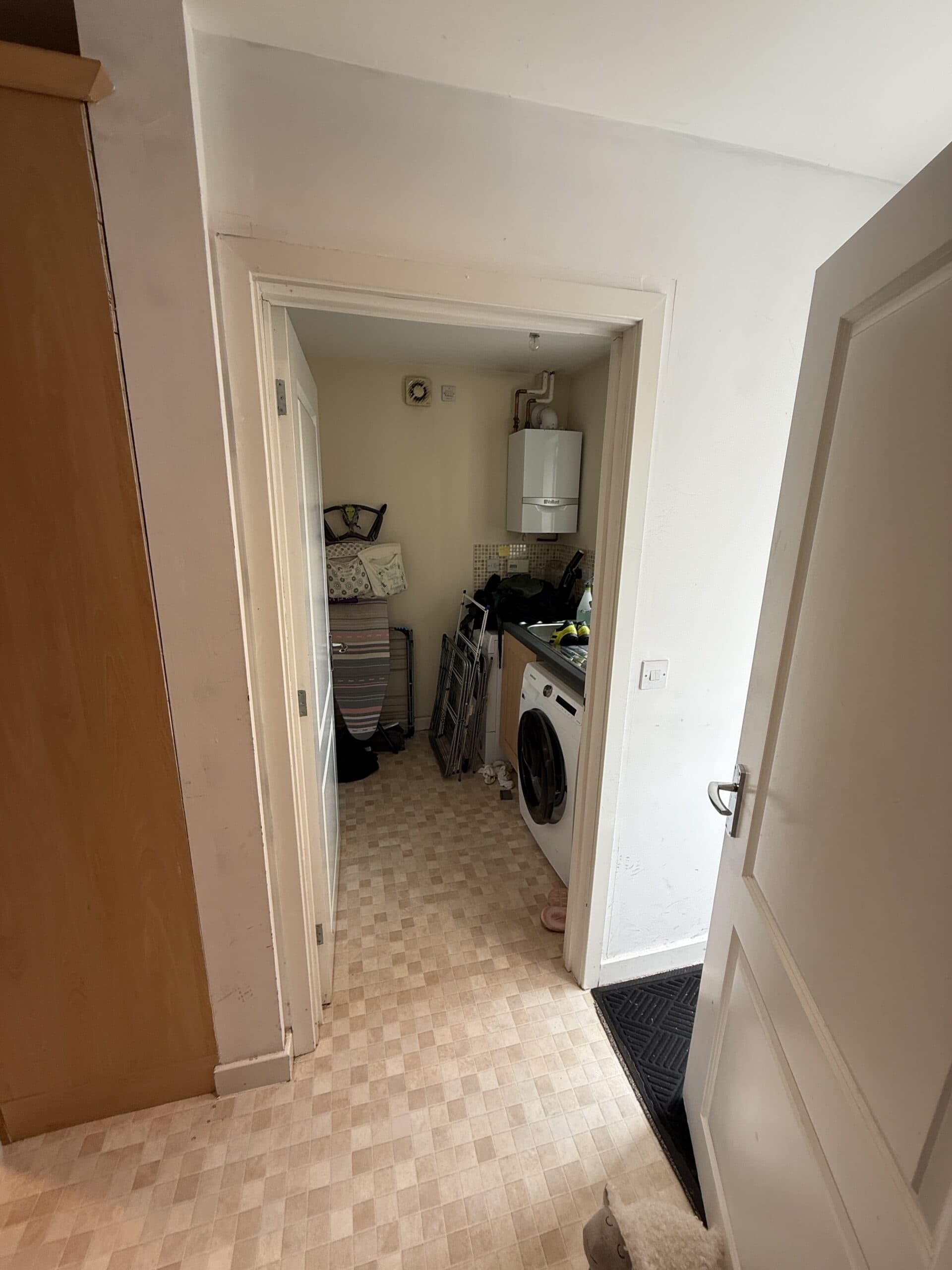 A view through an open door into a small utility room with a washing machine, stacked laundry baskets, shelves, a boiler on the wall, and various household items. The floor has a tiled pattern.