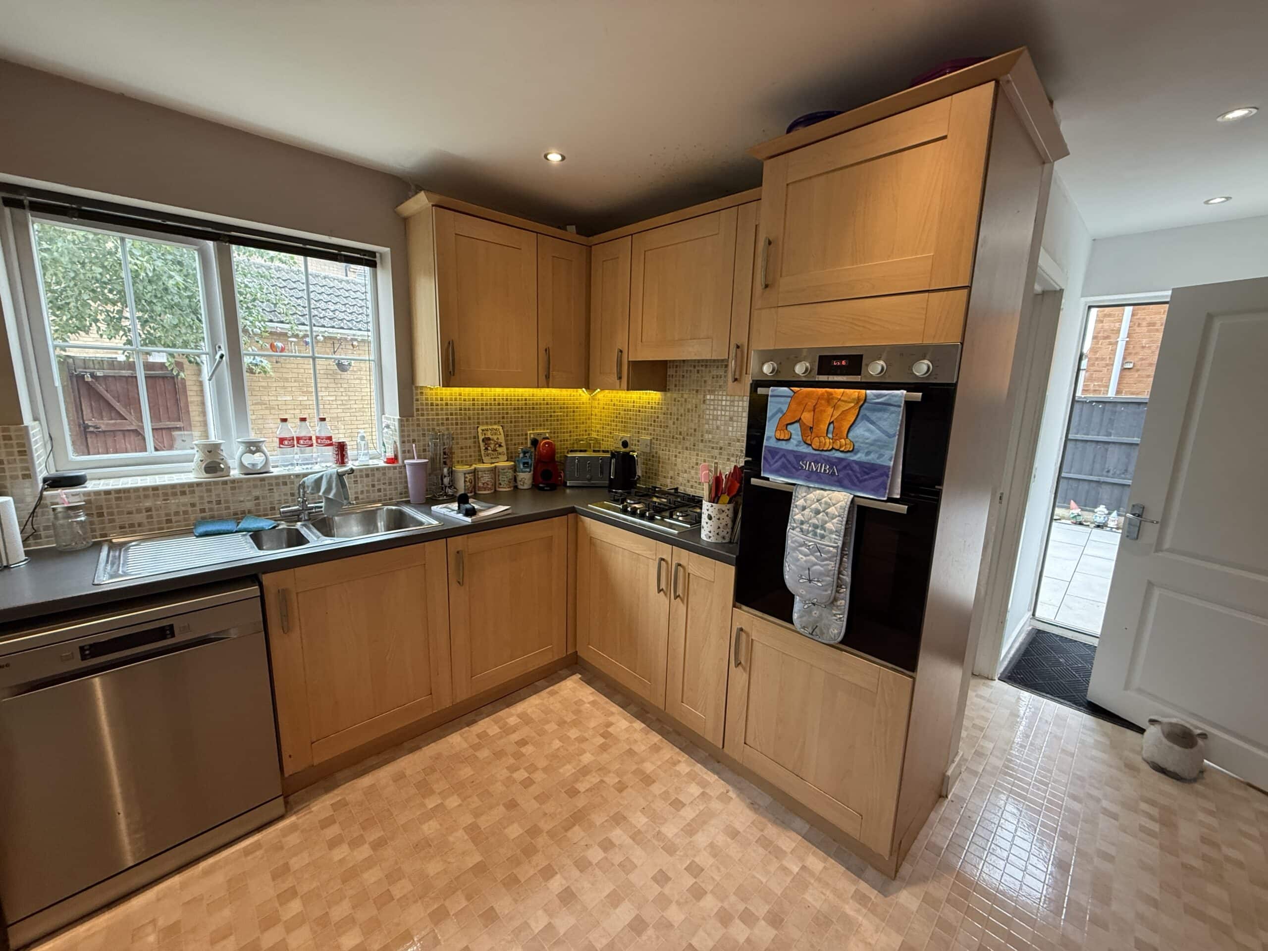 A kitchen with light wood cupboards, a built-in oven, hob, and dishwasher. The worktops hold various appliances. A tea towel and oven glove hang on the oven. Light tiles cover the floor. A window and door provide natural light.