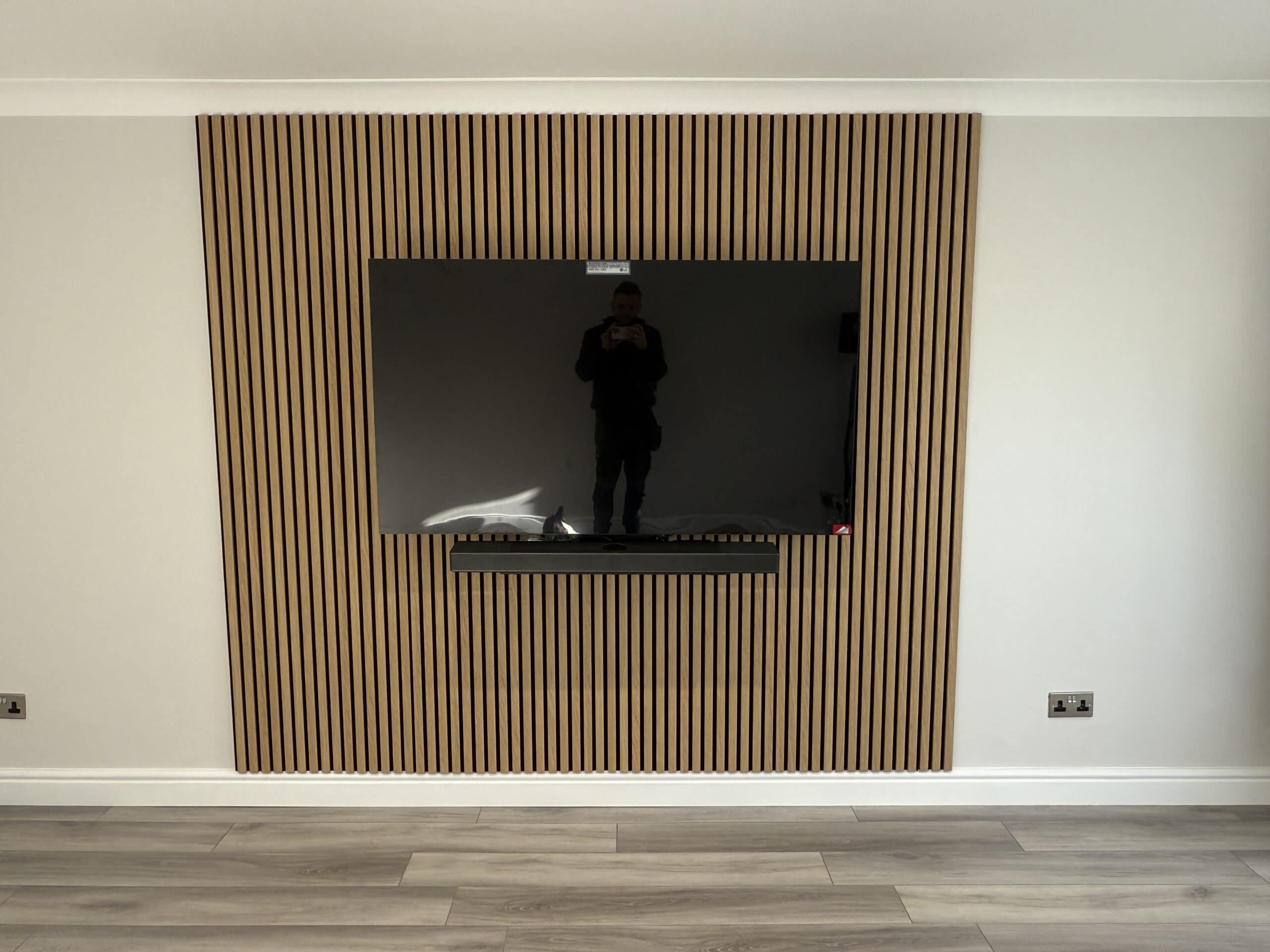 A flat-screen TV is mounted on a wooden slat feature wall in a modern, minimalist room with light-coloured flooring. A person is reflected in the TV screen whilst taking the photo.