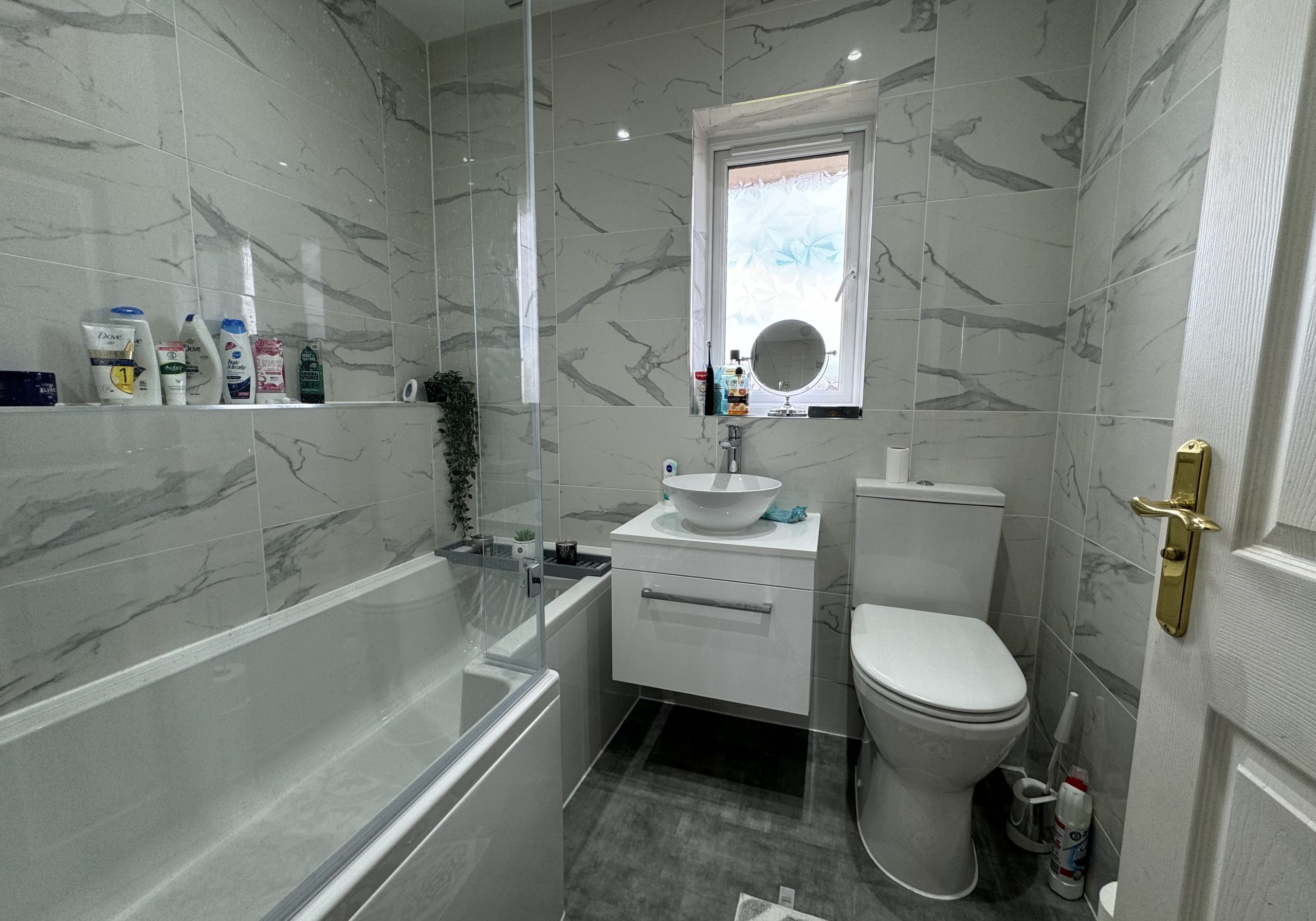 Modern bathroom with marble-patterned tiles, a bathtub with shower, white basin on a vanity unit, round mirror, toilet, and toiletries. A window lets in natural light—perfect inspiration for your next kitchen and bathroom showroom design. Modern bathroom with marble-patterned tiles, a bathtub with shower, white basin on a vanity unit, round mirror, toilet, and toiletries. A window lets in natural light—perfect inspiration for your next kitchen and bathroom showroom design.