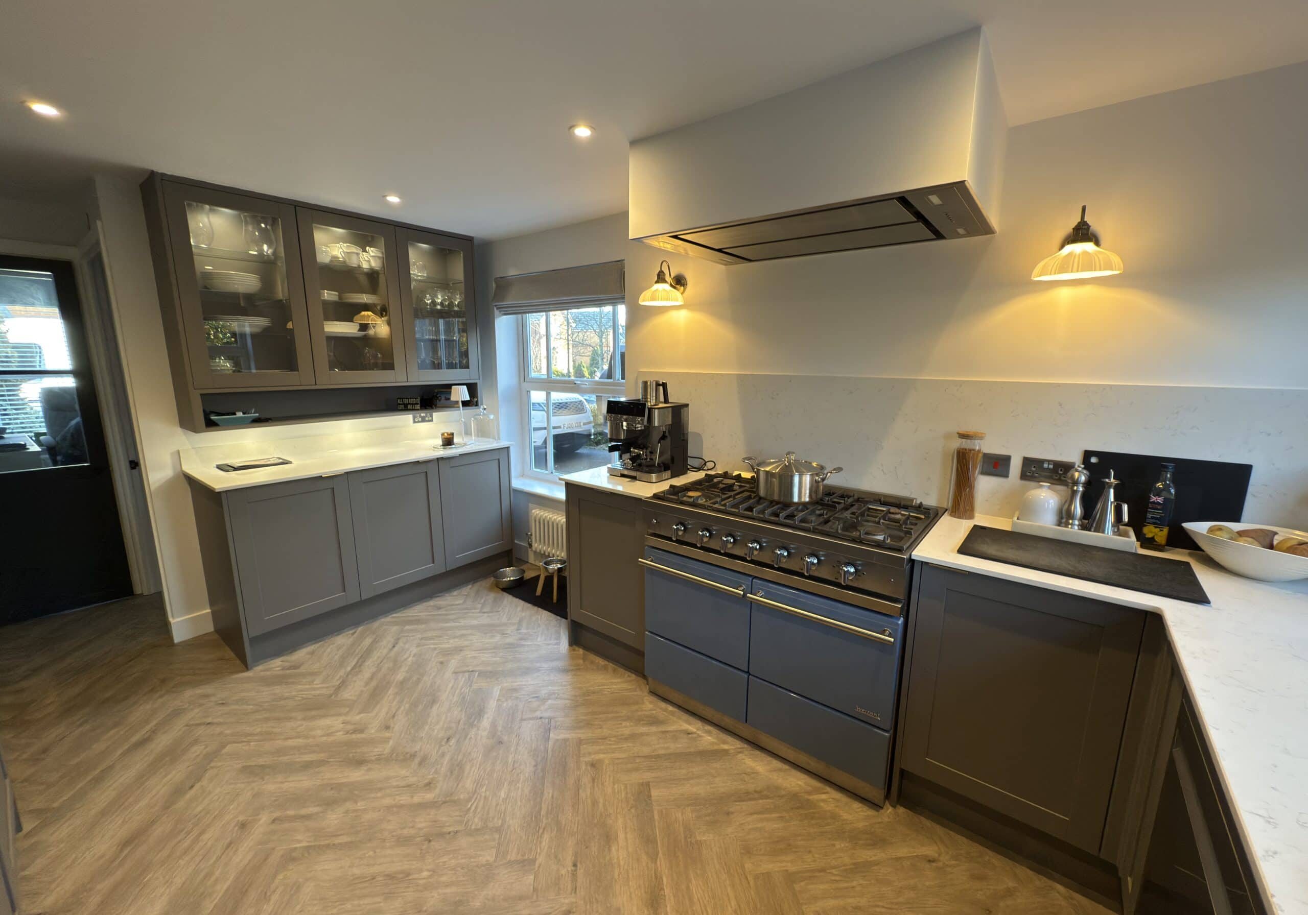 Modern kitchen and bathroom showroom design featuring grey cupboards, a large cooker, white worktops, glass-fronted cabinets, wooden herringbone flooring, and under-cupboard lighting; window and door let in natural light.
