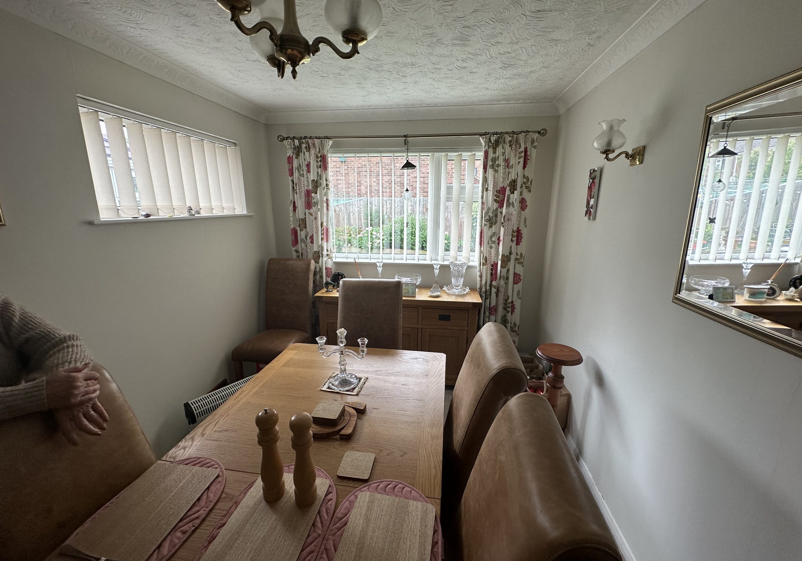A dining room with a wooden table set for four, brown leather chairs, placemats, and pepper mills offers a warm space. Large windows with blinds and floral curtains echo thoughtful design found in a kitchen and bathroom showroom. A person's arm rests at the table's edge.