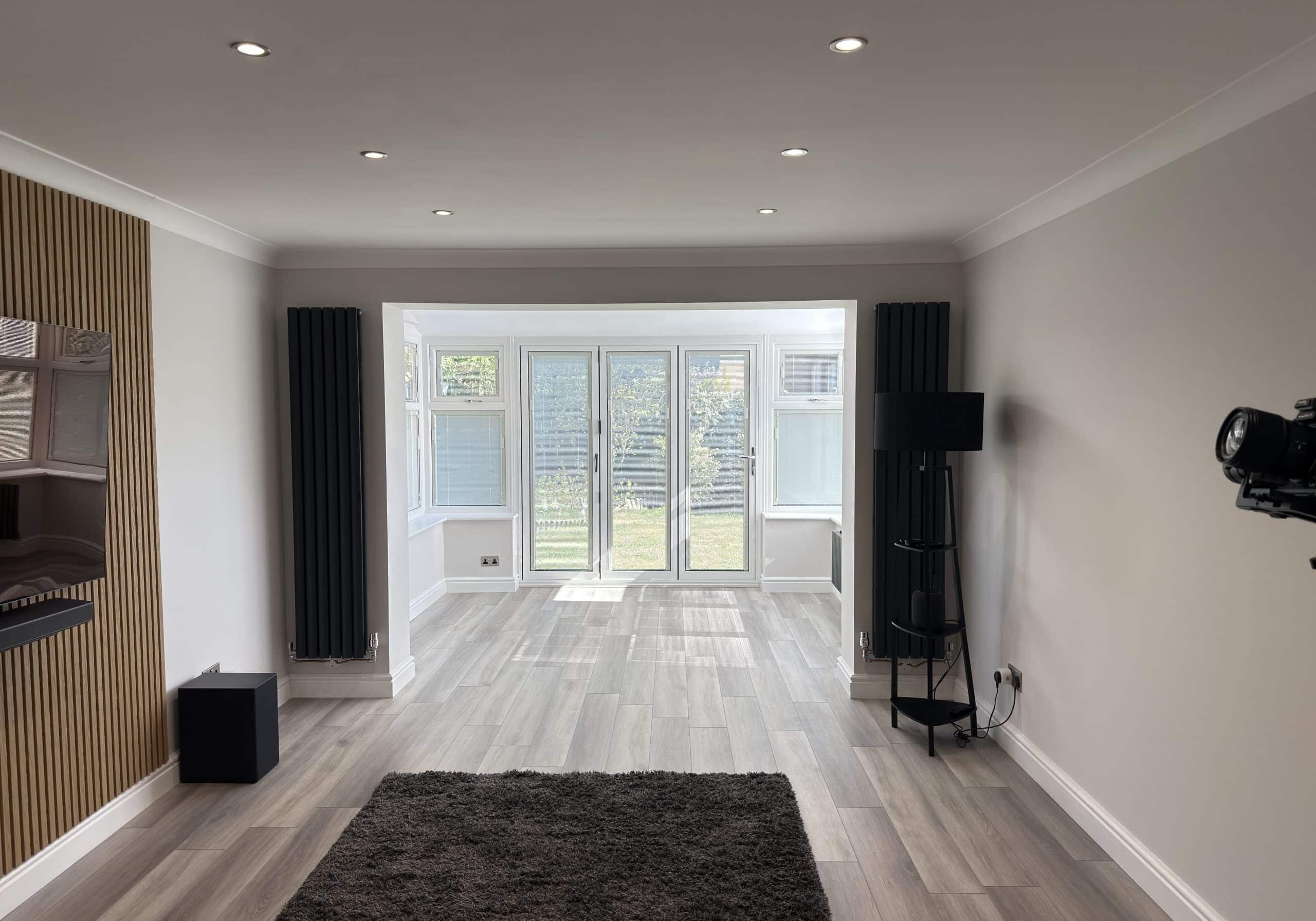 A modern, unfurnished lounge with light grey walls, wooden flooring, and recessed ceiling lights. Perfect for showcasing kitchen and bathroom showroom installation supply design ideas, it features large windows and glass doors opening to a green garden view.