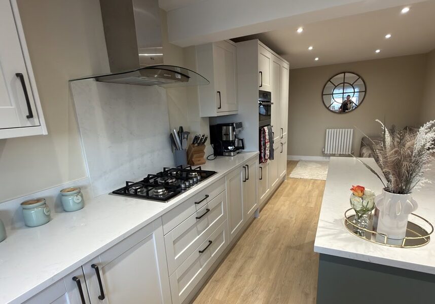 Modern kitchen with white cupboards, gas cooker, and countertop appliances. Light wood floor, neutral walls, decorative vases, and a tray with dried flowers on the island. Round mirror and framed picture in the dining area.