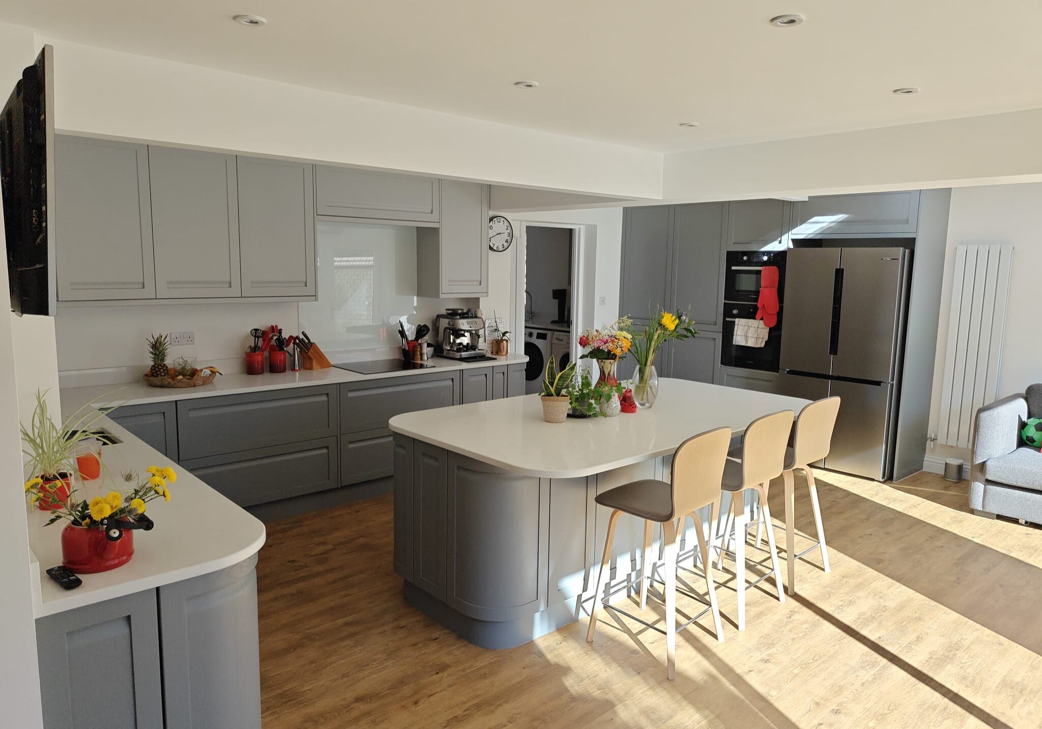 Modern kitchen with grey cupboards, white worktops, and wooden flooring. There are three beige bar stools at an island, fresh flowers, fruit, and kitchen appliances visible, with sunlight streaming in through large windows.