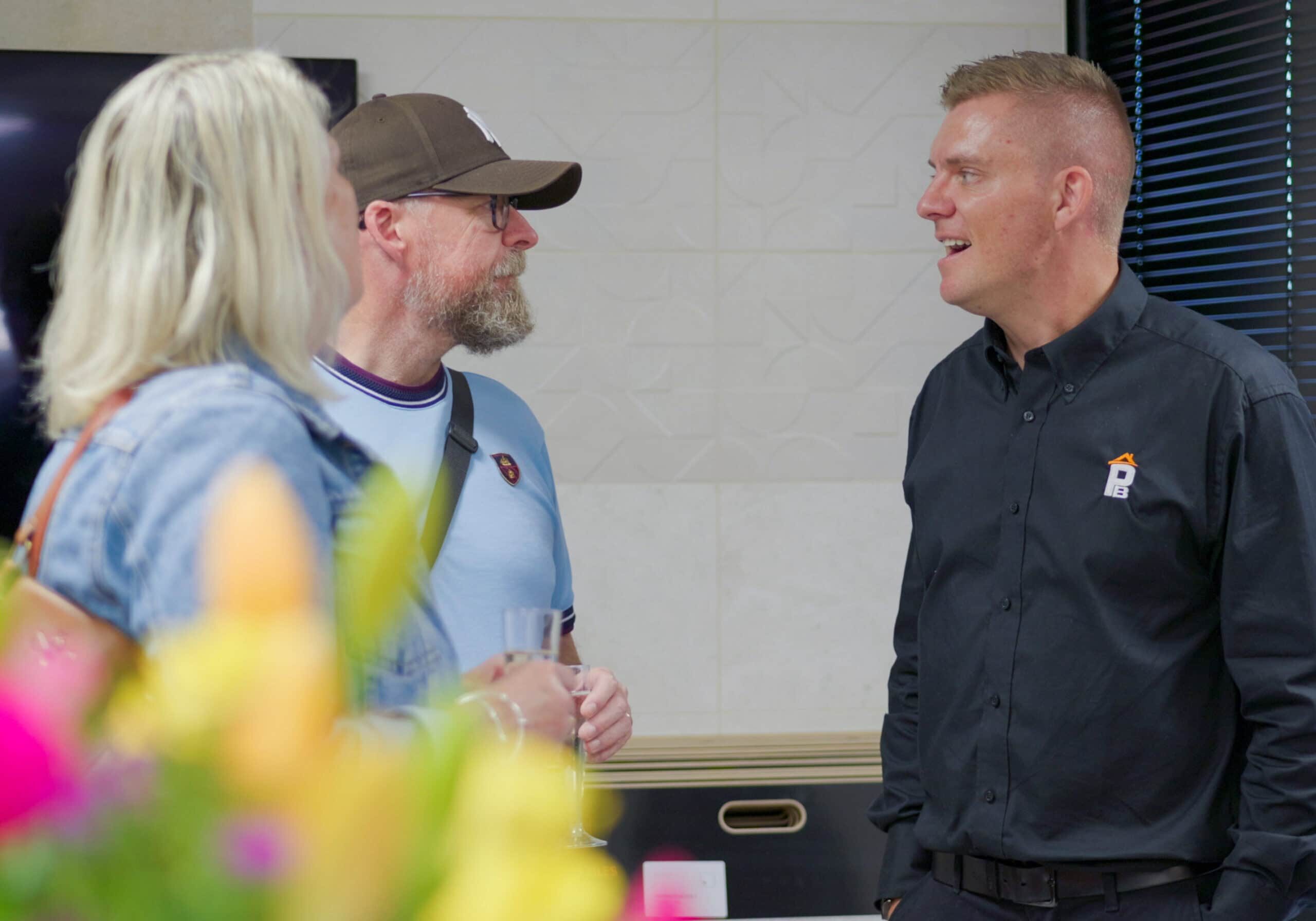 Three adults are talking indoors. A man in a black shirt speaks to a man wearing a cap and glasses, who stands beside a woman with blonde hair. Colourful blurred flowers are in the foreground.
