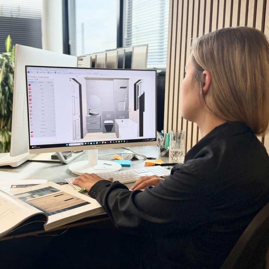 A woman sits at a desk, working on a computer with a bathroom design programme open. An open magazine and various office supplies are on the desk.